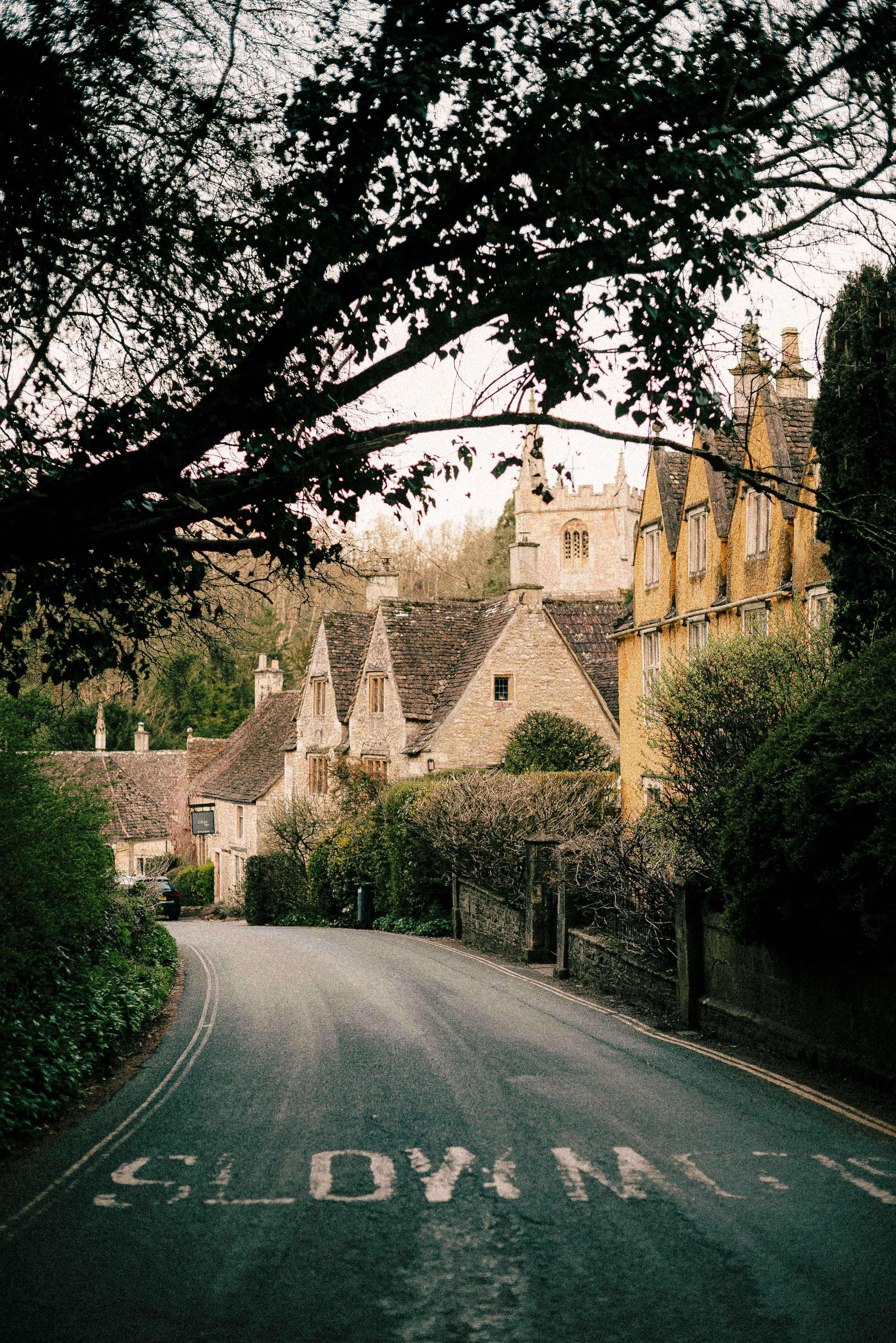 A winding village road leads through traditional Cotswold stone buildings and cottages in rural England.