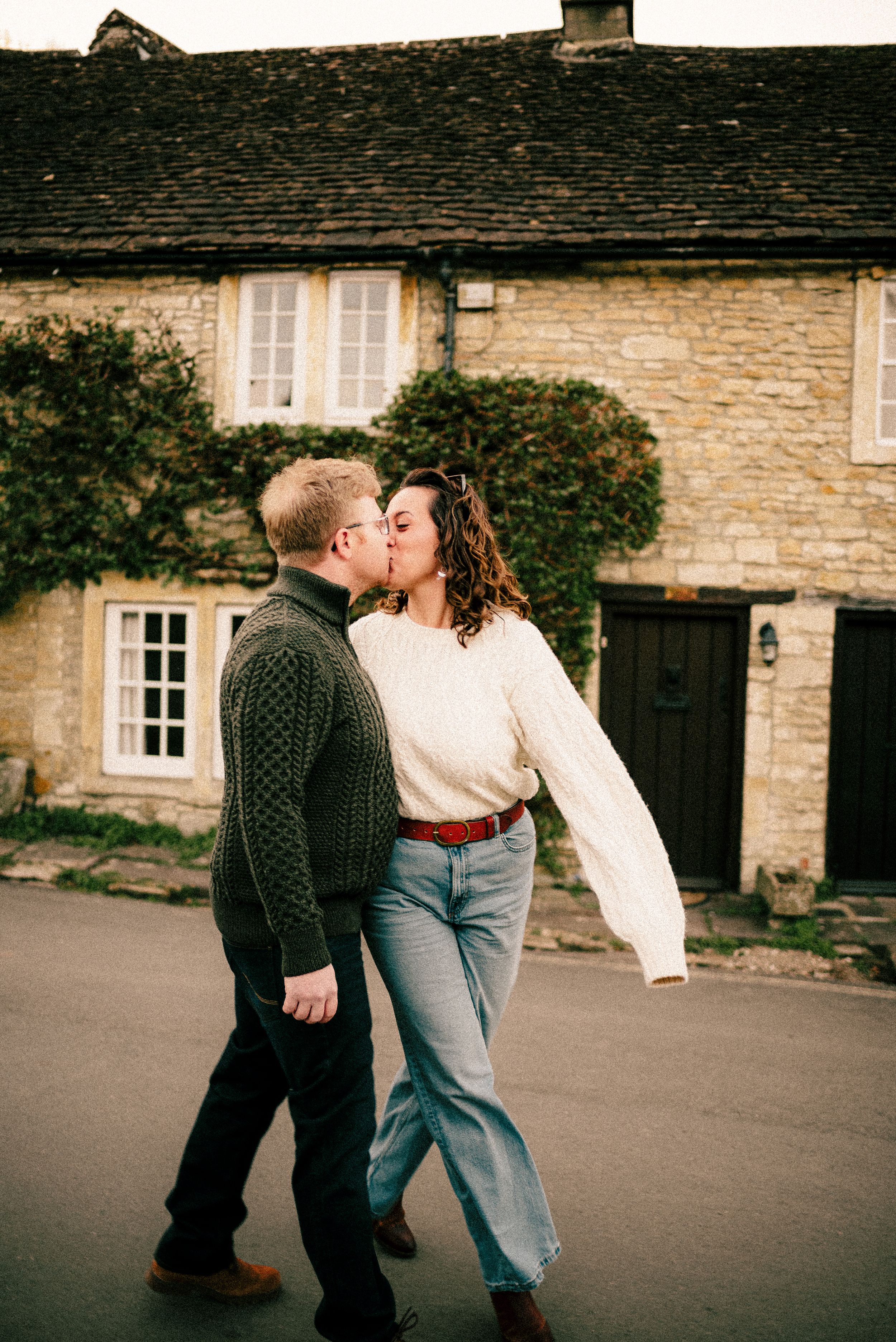 A couple in casual attire sharing a romantic moment against a rustic stone building backdrop.