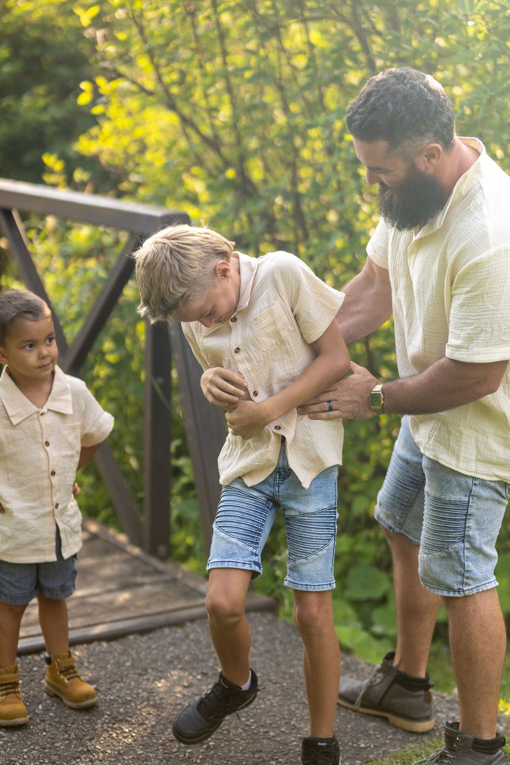 A father tickling his son in order to get him to smile and the younger bother watching