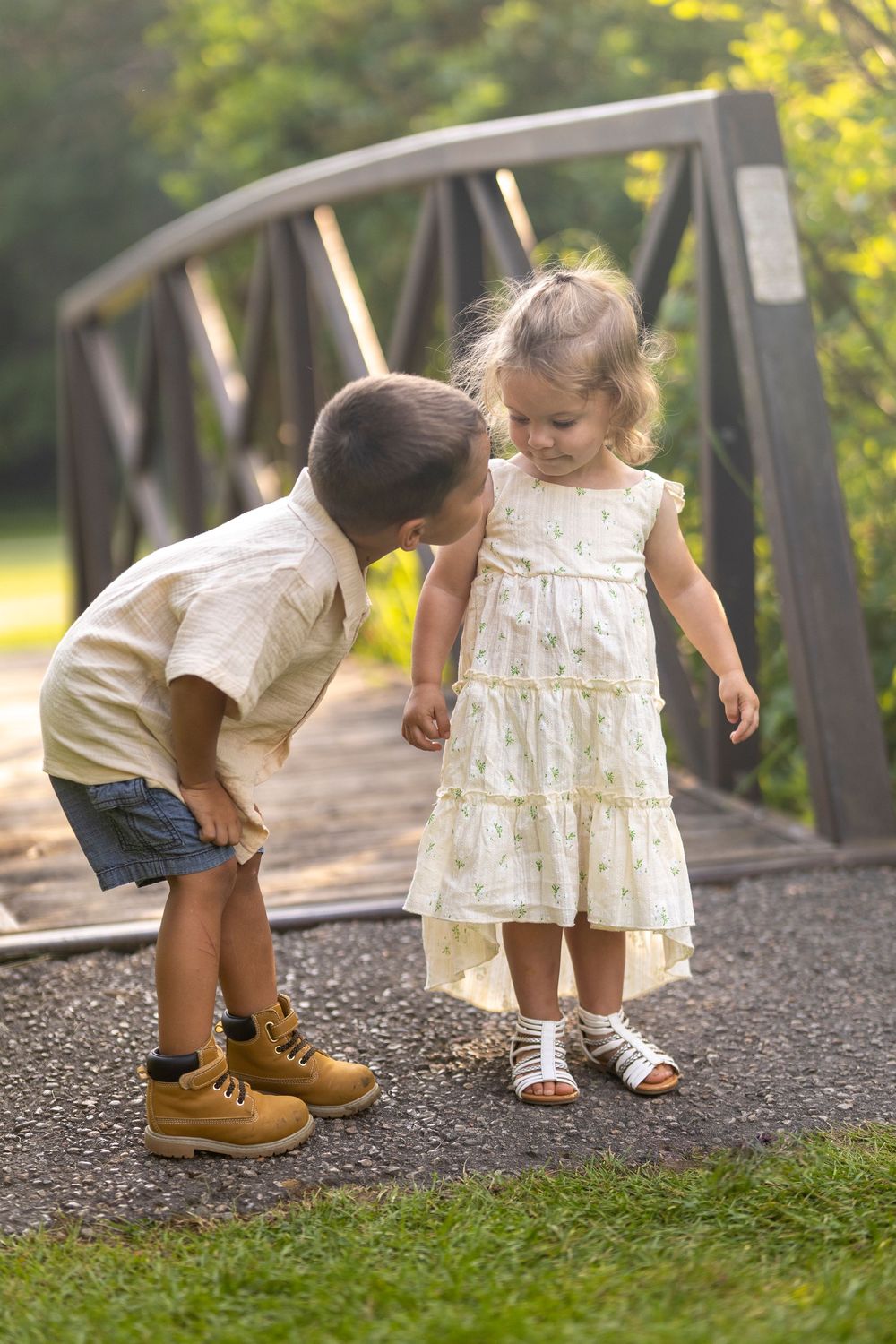 A young brother and sister near a bridge where the brother is leaning down to look his sister in the face