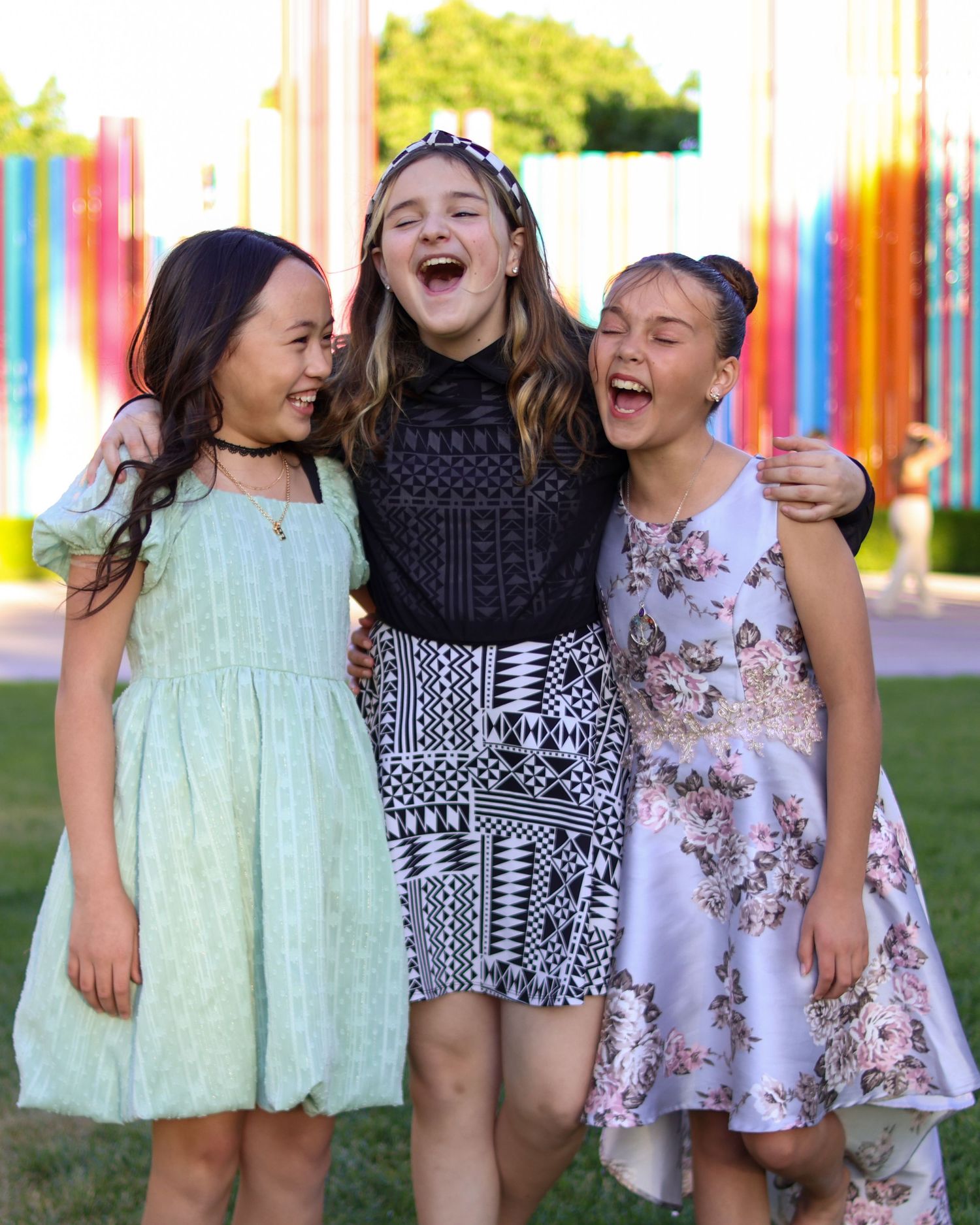 Three friends in summer dresses share a joyful laugh against a colorful outdoor backdrop.