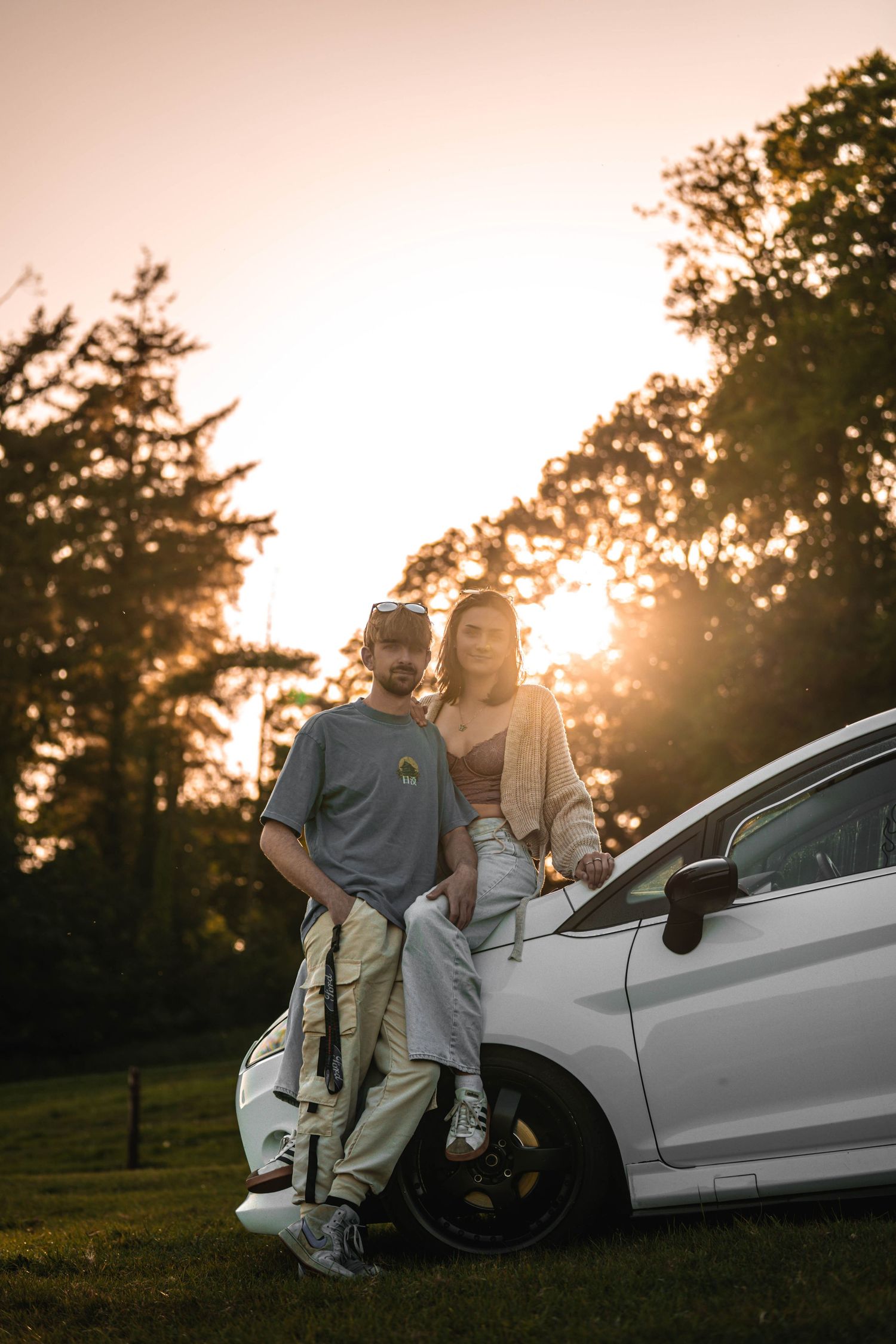 Couple leaning against white car during golden hour sunset with trees in background.