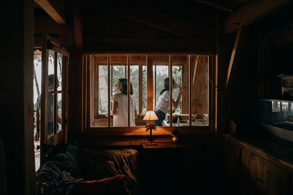 Cozy cabin interior with wooden walls and window frames lit by a warm table lamp.