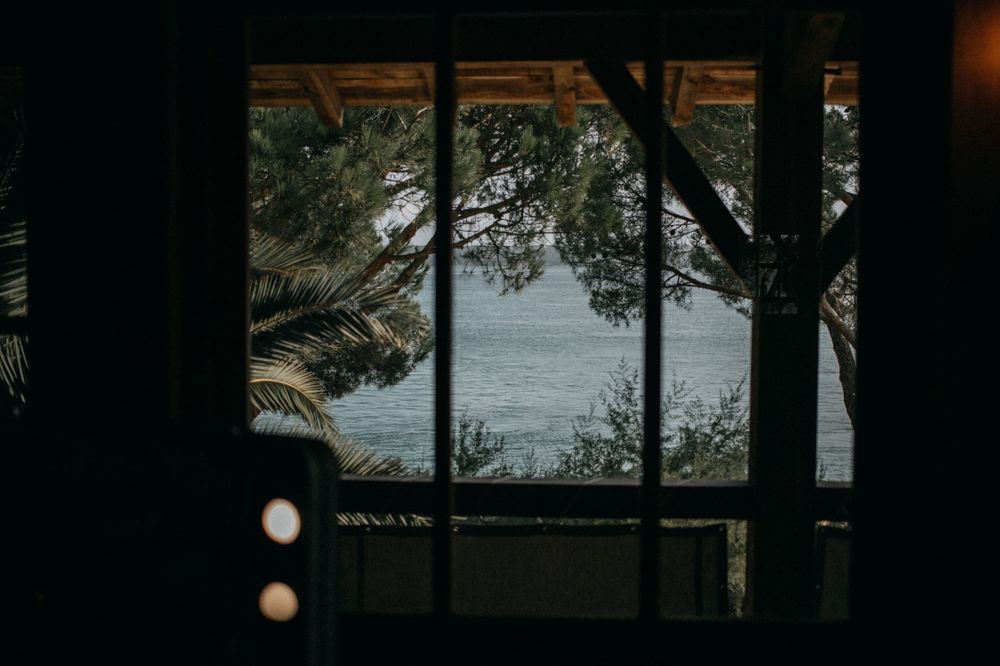 Ocean view through dark-framed windows of a coastal cabin with palm trees silhouetted against the water.