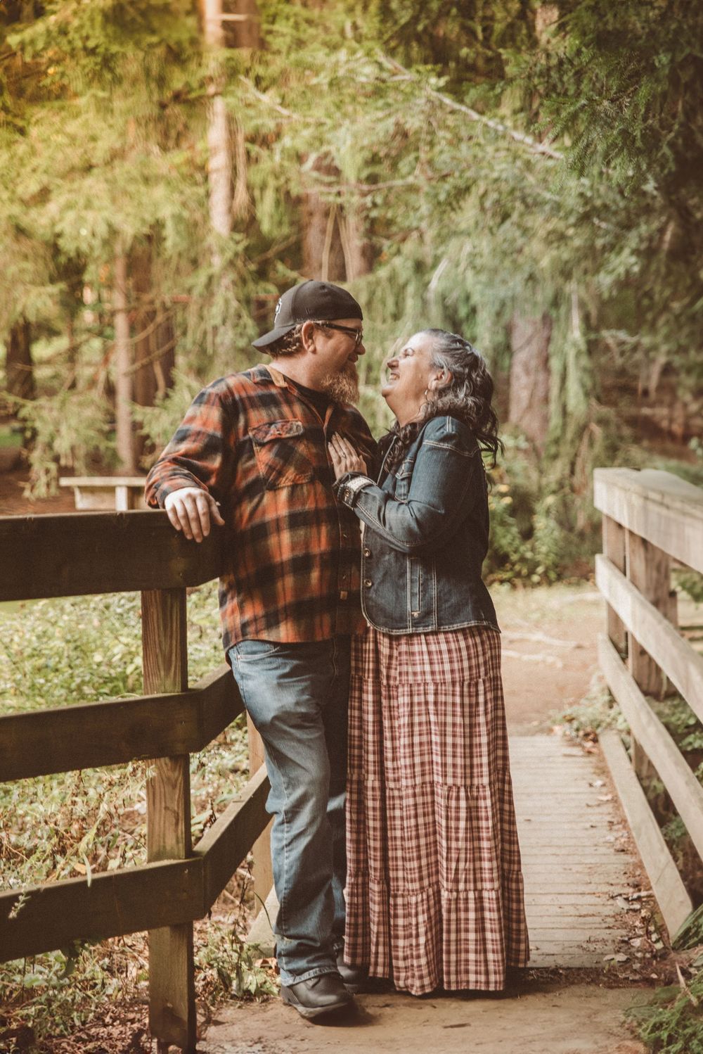 A couple smiling at each other in a forested area on a small wooden bridge.