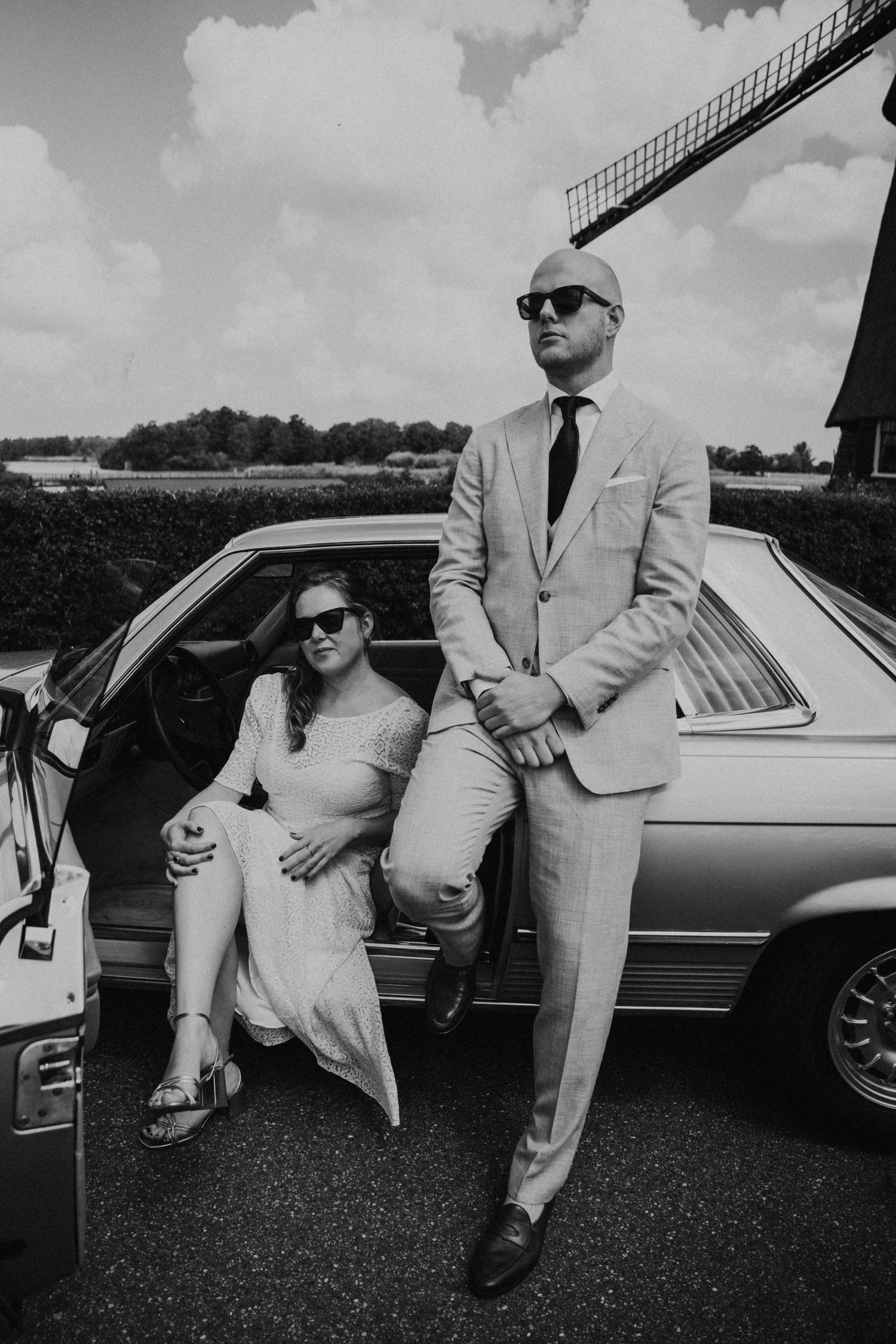 Stylish couple poses with vintage car wearing sunglasses and formal attire.
