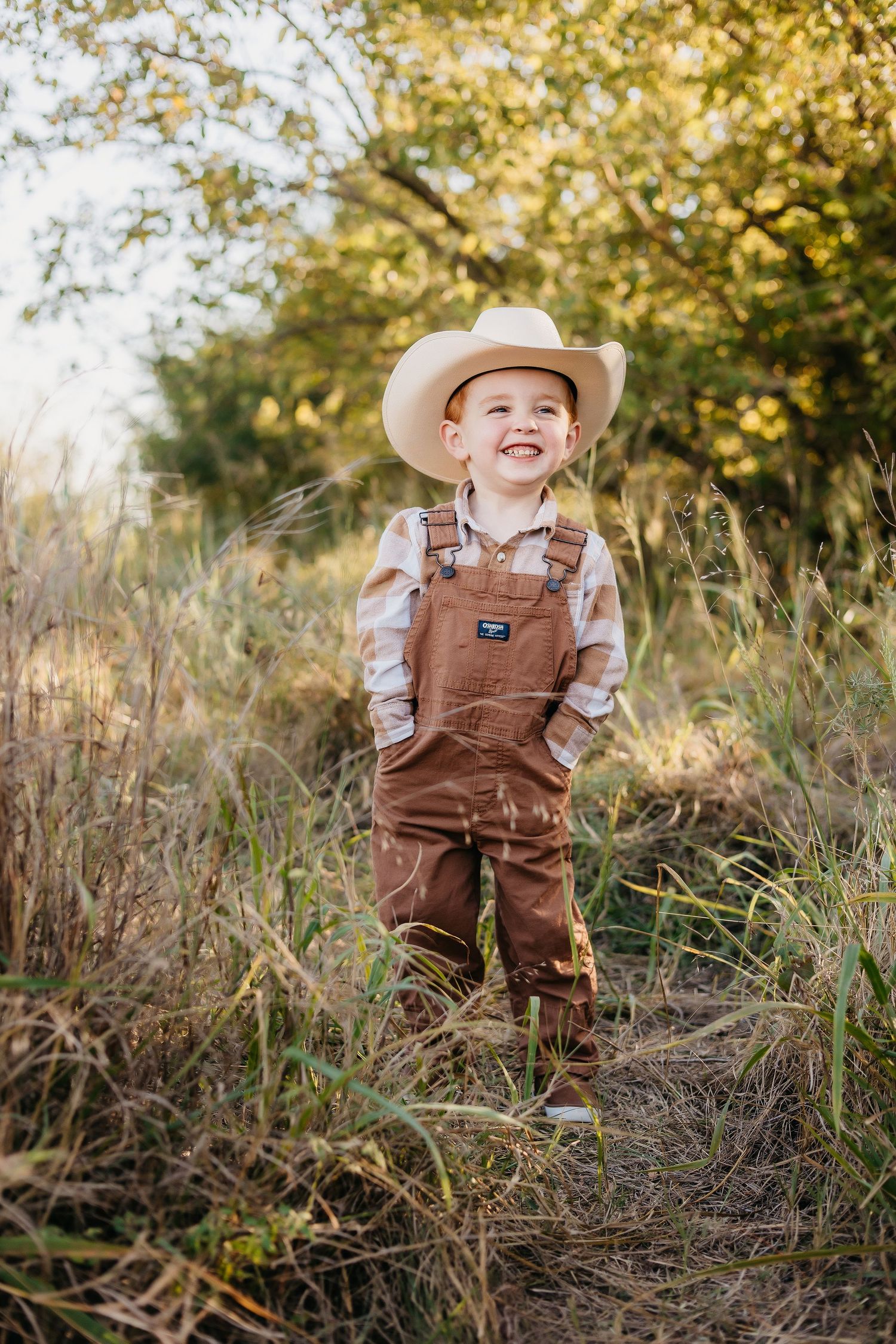 Small child wearing brown overalls and cowboy hat playing in tall autumn grass.