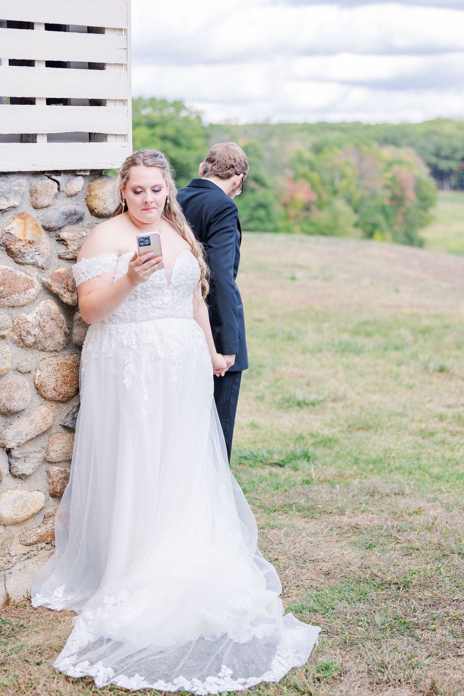 A bride and groom stand back to back checking their phones against a rustic stone wall.