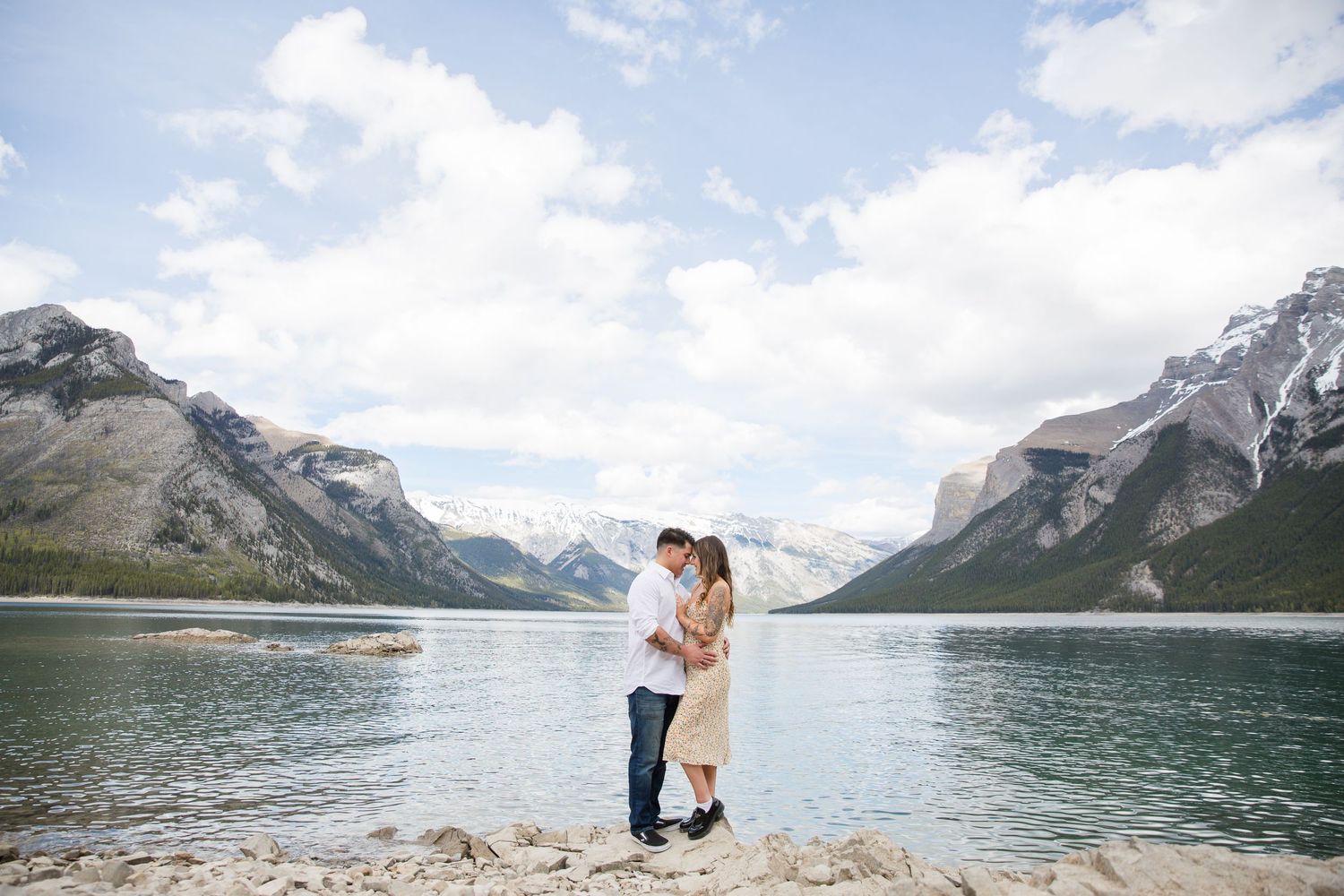 Couple embracing on shore of mountain lake with snow-capped peaks in background.