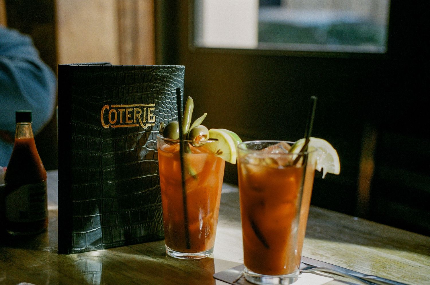 Two refreshing iced cocktails with citrus garnishes sit next to a menu on a wooden table in a dimly lit bar.
