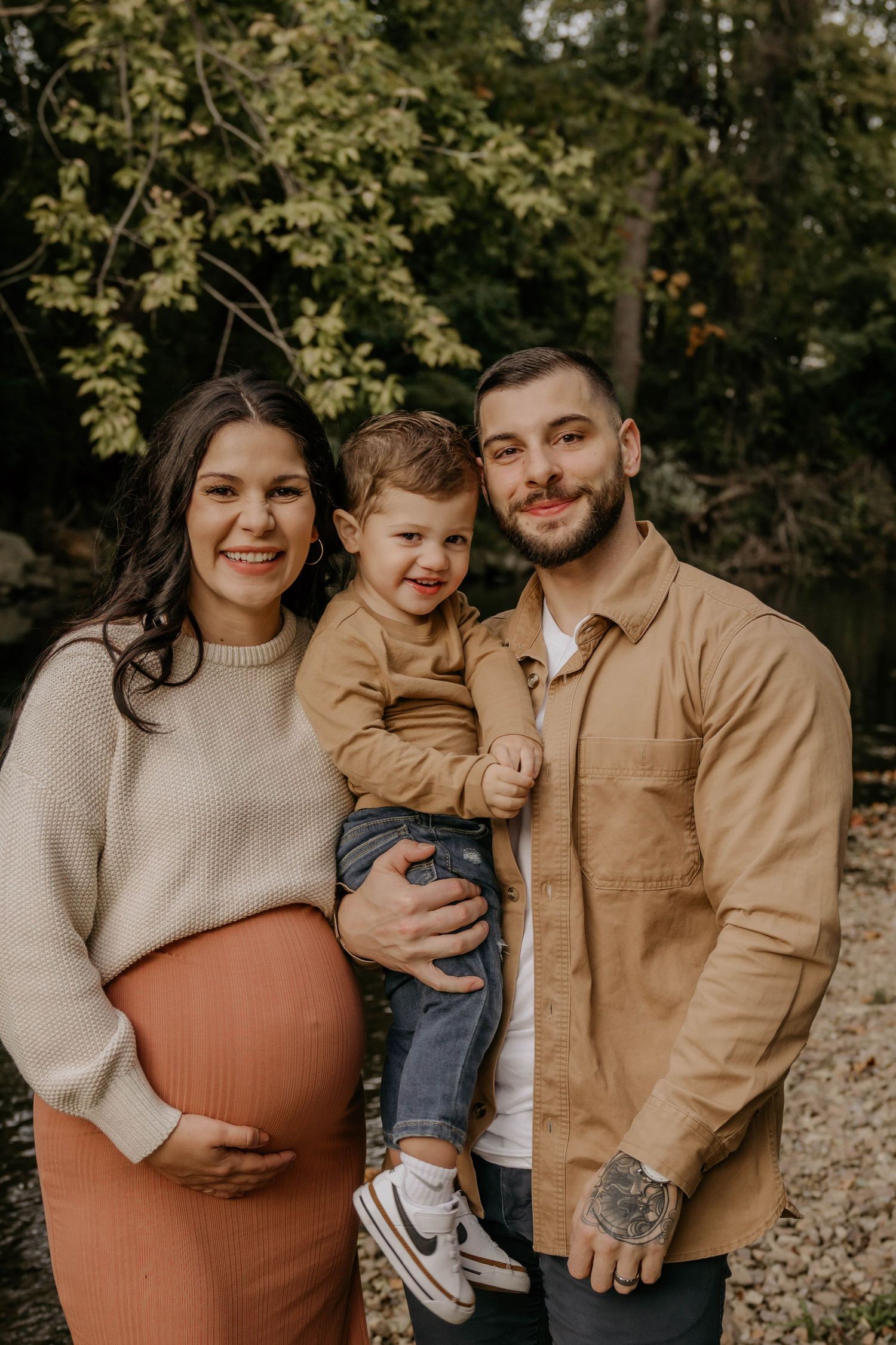 Pregnant woman in cream sweater poses with toddler son and husband outdoors in autumn setting.