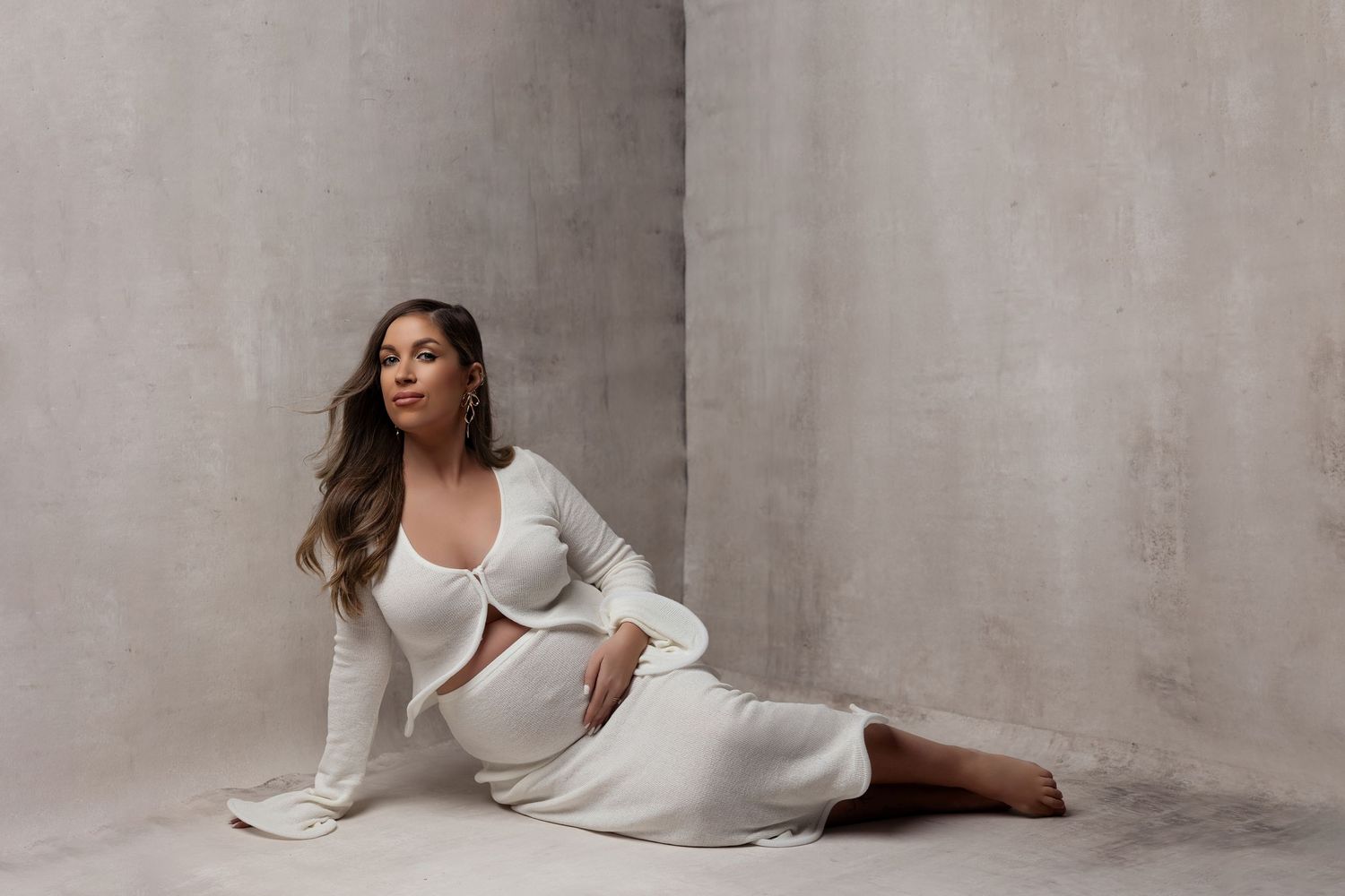 Fashion model in white pantsuit and crop top poses gracefully on floor against textured gray backdrop in studio setting.