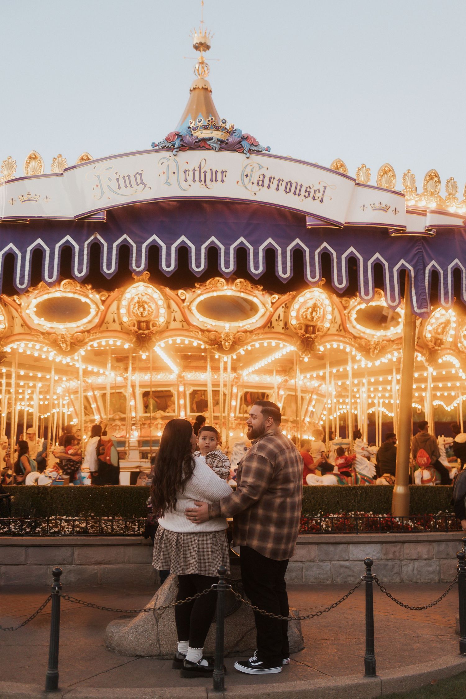 A couple stands in front of the illuminated King Arthur Carousel at night with glowing lights creating a magical atmosphere.