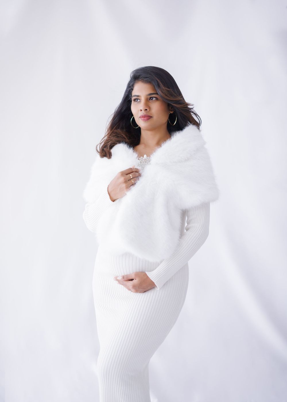 A maternity portrait in an elegant white knit dress with plush fur wrap against a light backdrop.