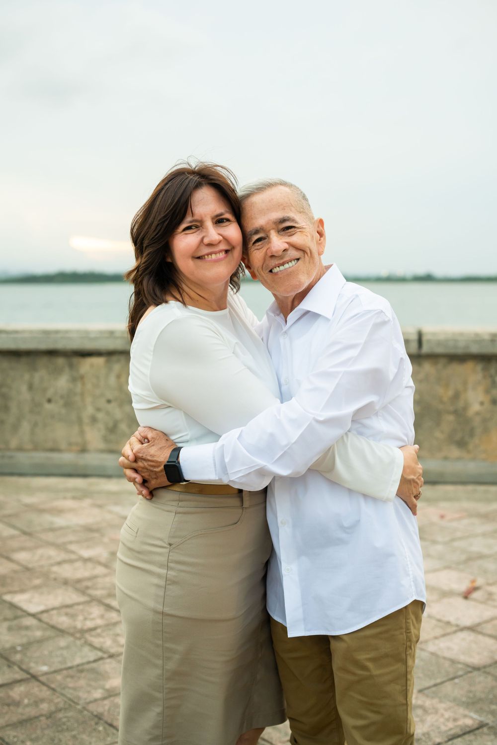 Couple in matching white shirts share loving embrace by waterfront.