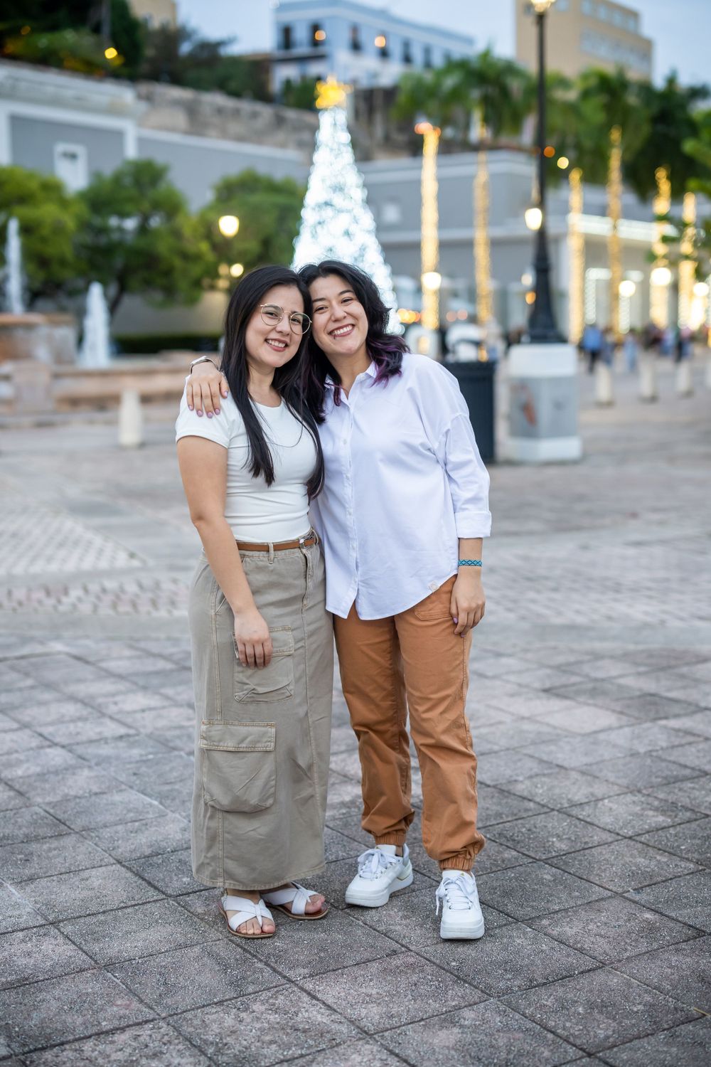 Two people in casual outfits stand together smiling with a decorated Christmas tree in the background.