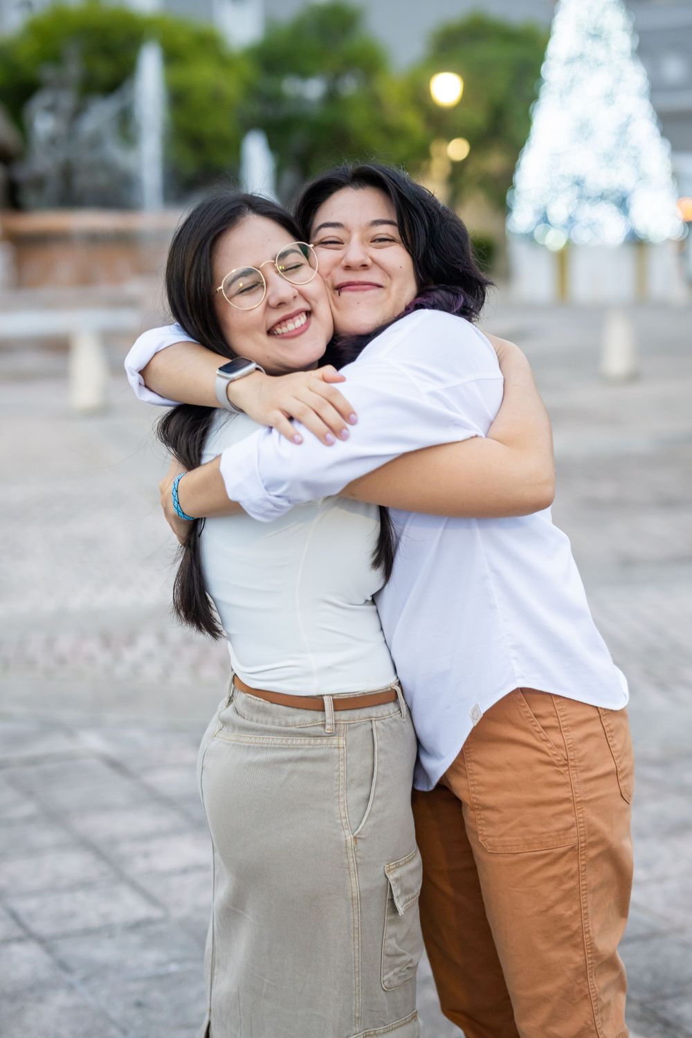 Two people share a tight hug and big smiles with holiday decorations visible behind them.