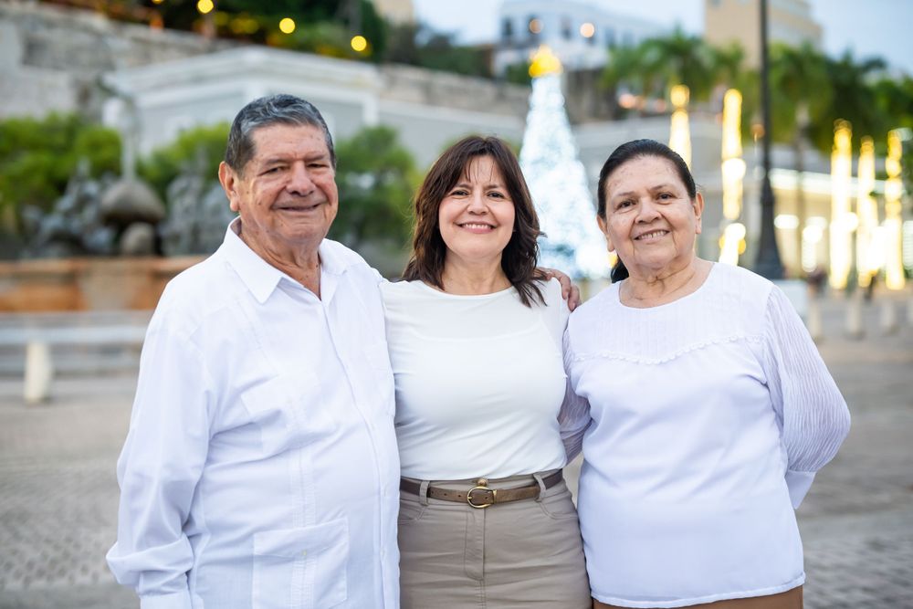 Three people in white shirts pose together outdoors with holiday decorations in the background.