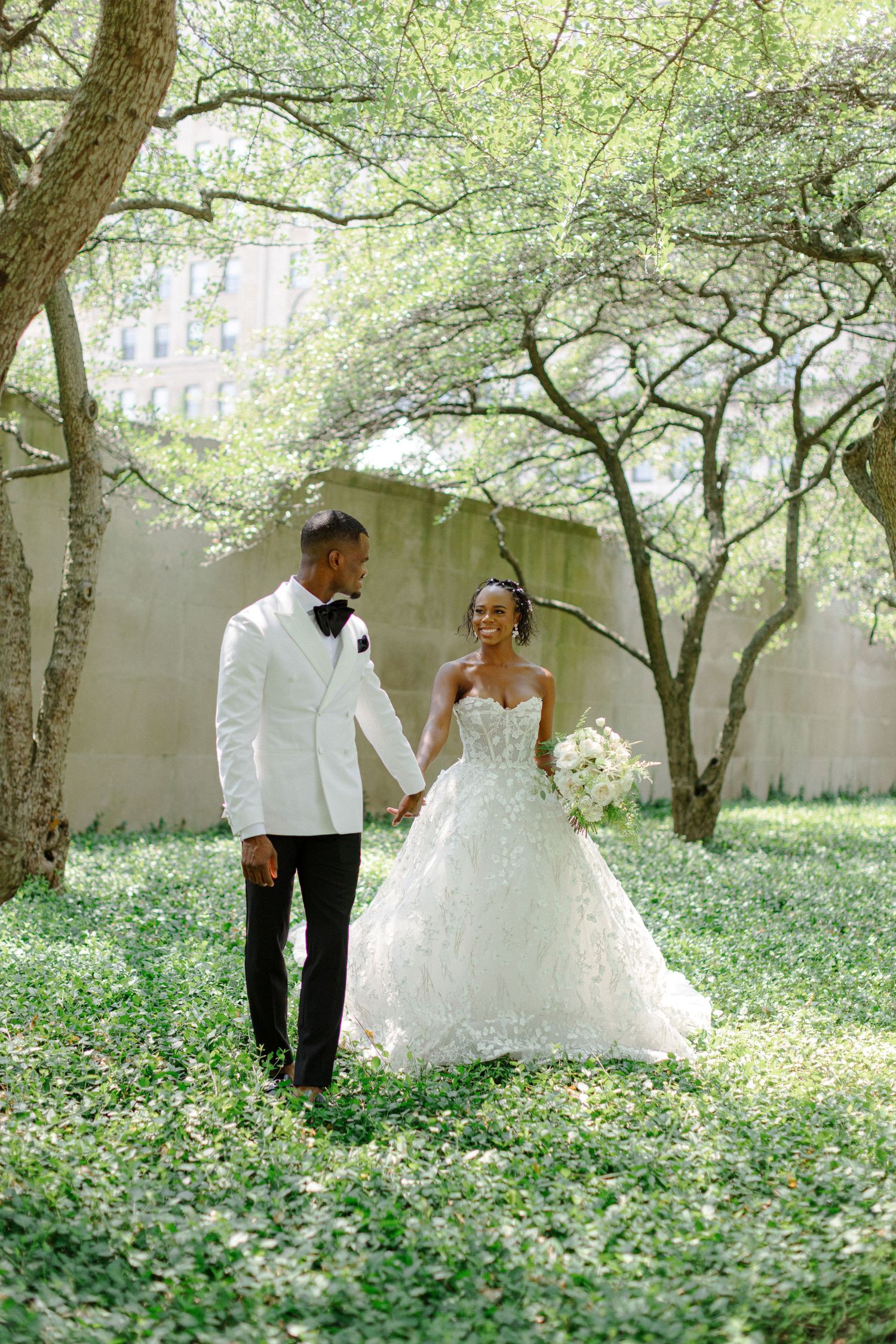 Newlyweds walk through lush garden setting in elegant white attire.