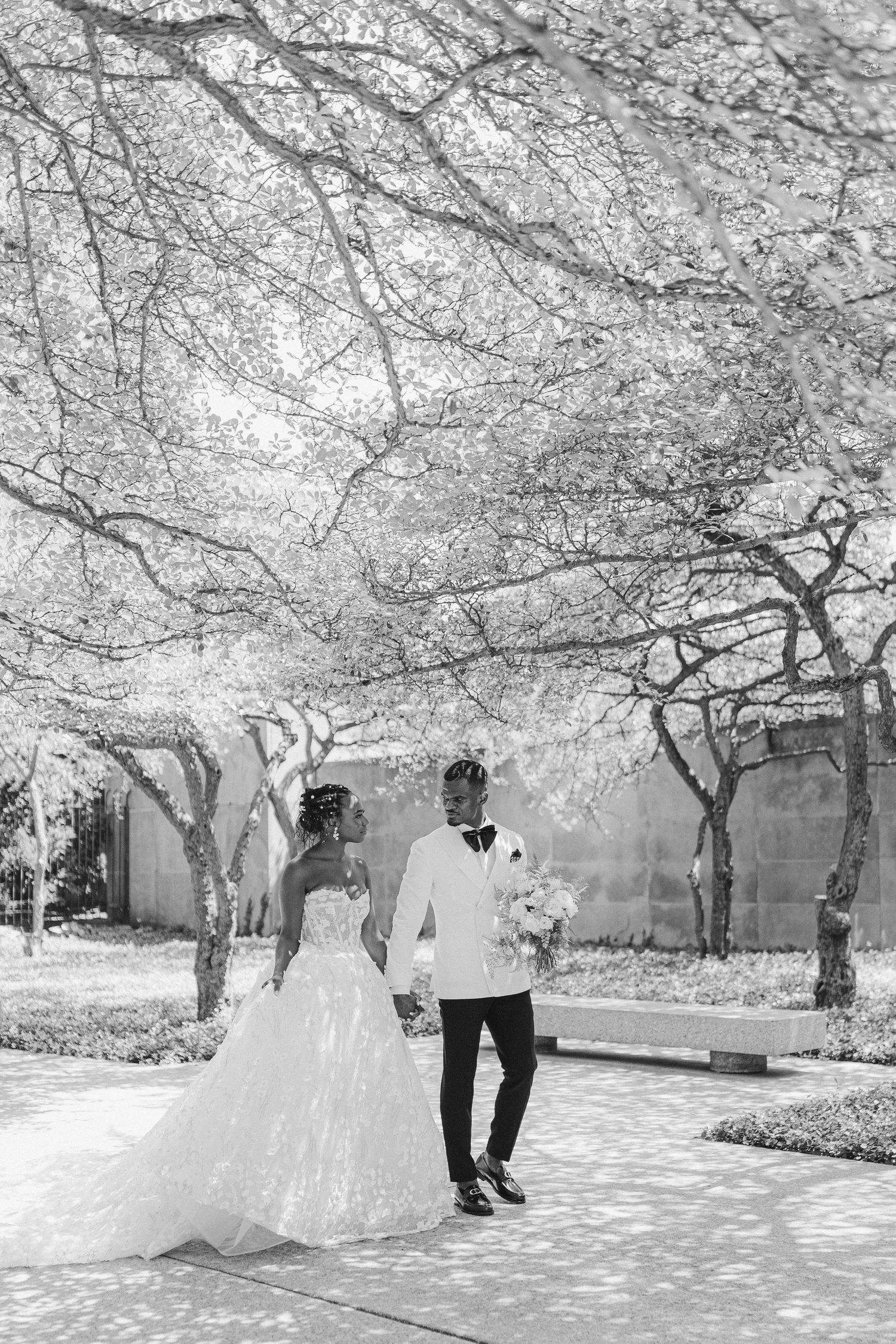 A black and white wedding photo of a couple walking beneath blooming cherry trees.