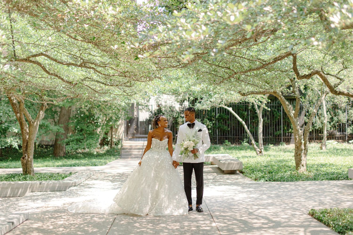 Newlyweds walk hand in hand through a sunlit garden pathway.