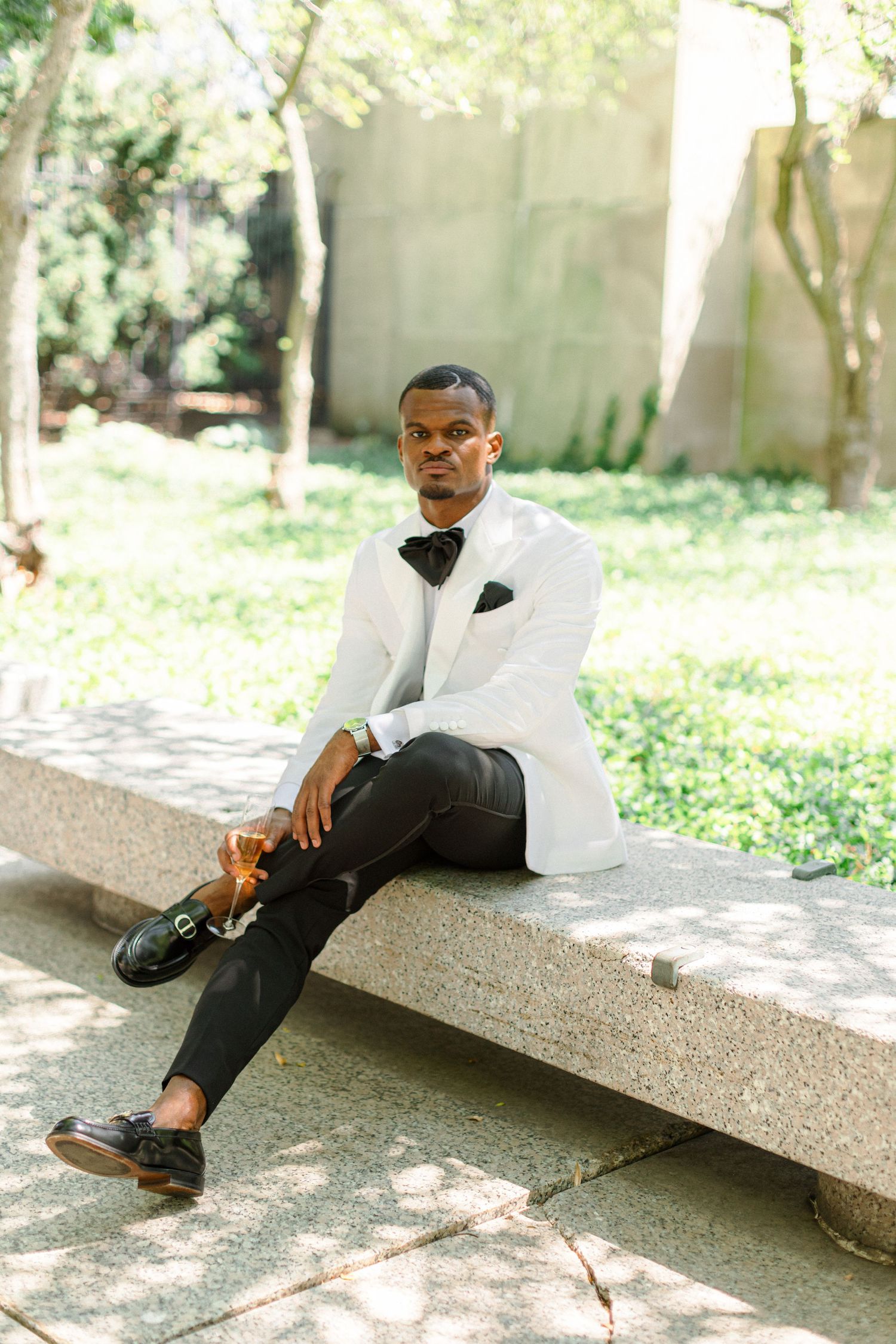 A formal portrait of someone in a crisp white tuxedo jacket seated on a stone bench.