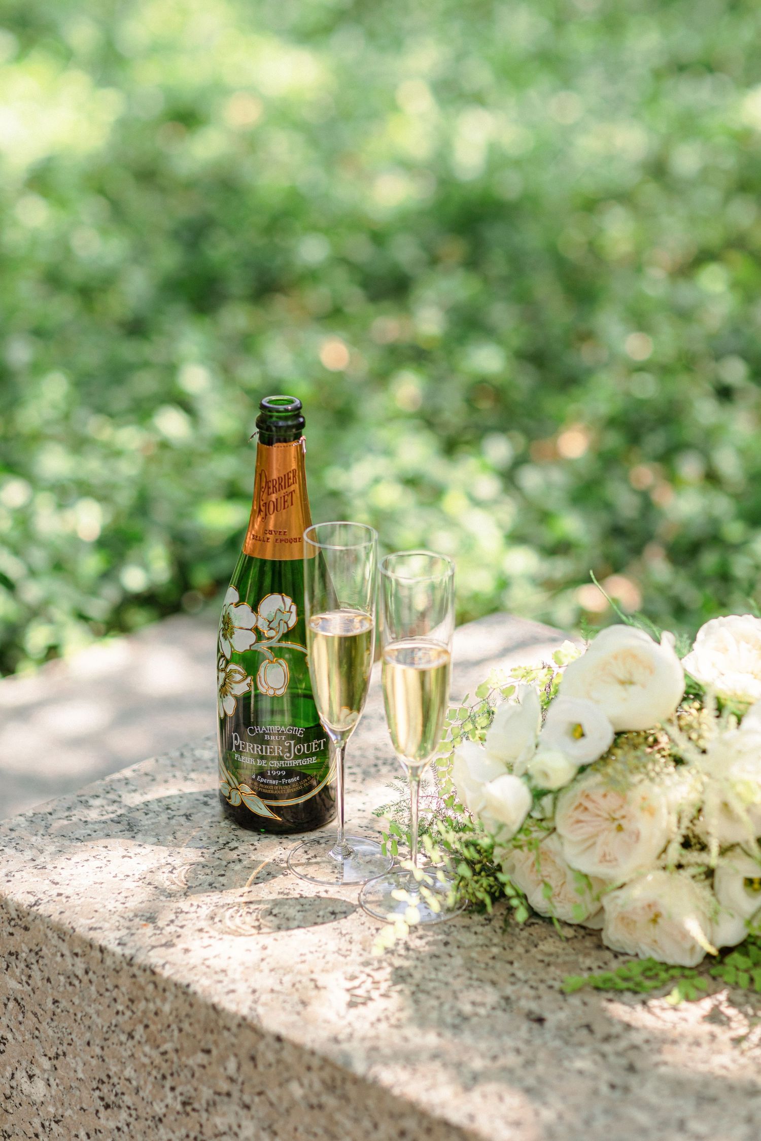 Champagne bottle and glasses next to white roses on a stone ledge in a garden setting.