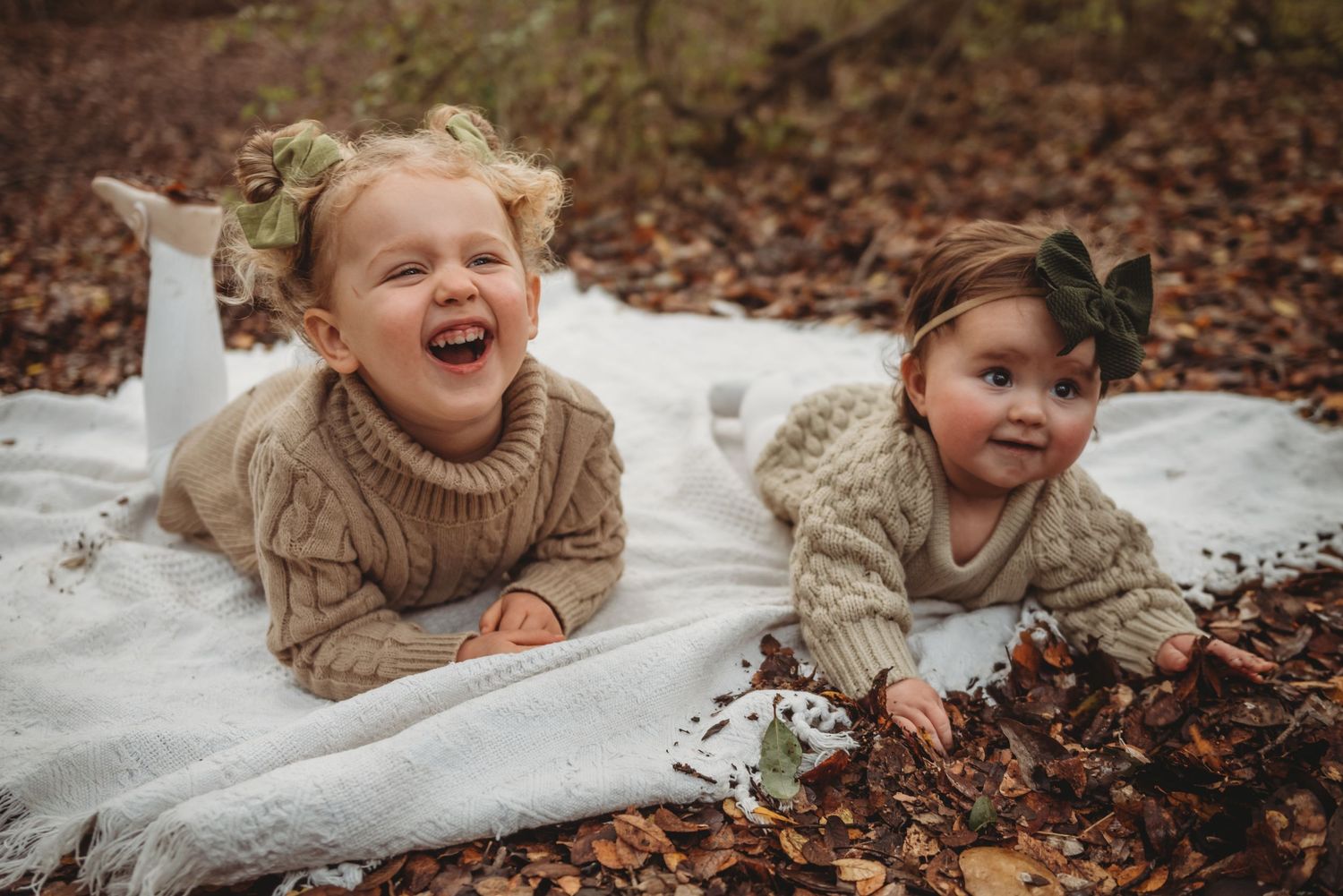 Two children laying on white blanket laughing and playing in fallen autumn leaves.