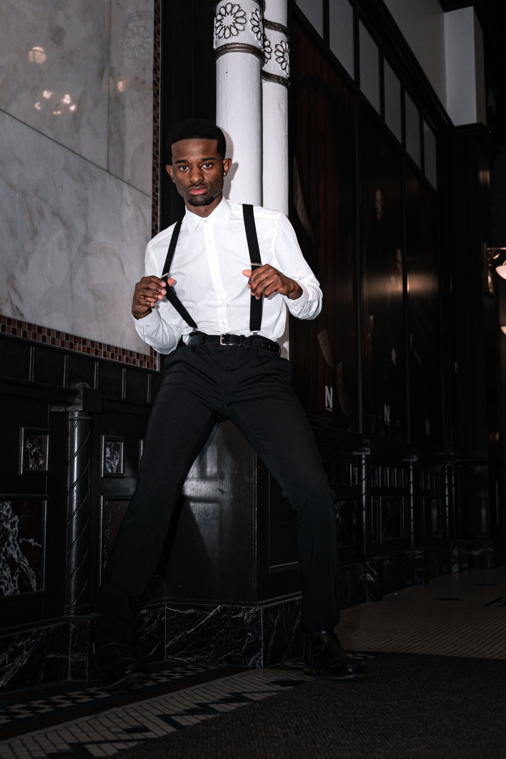 Stylish formal portrait with white shirt and suspenders against marble wall.