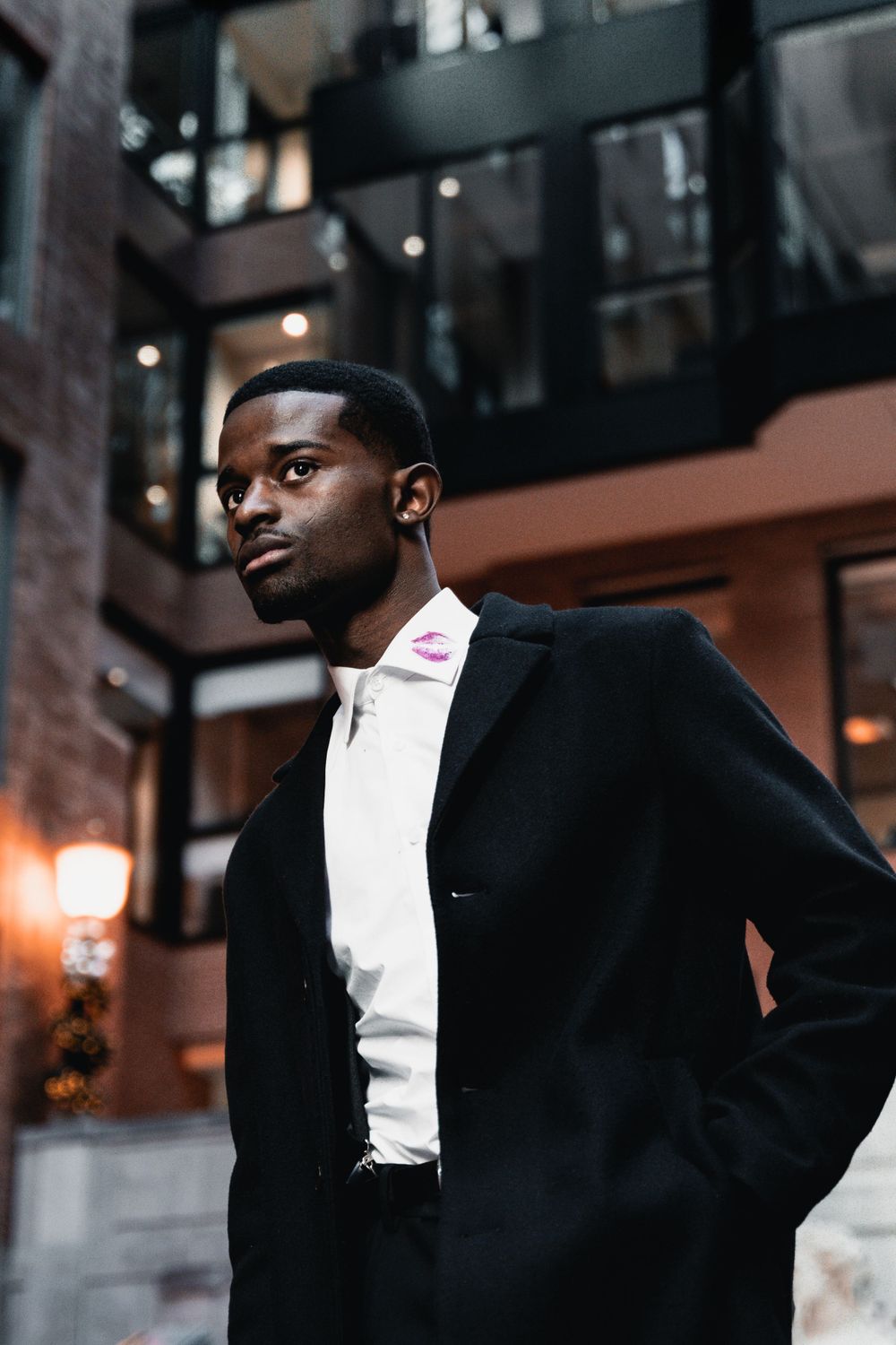 Stylish man in black suit and white shirt poses dramatically against urban building backdrop at dusk.