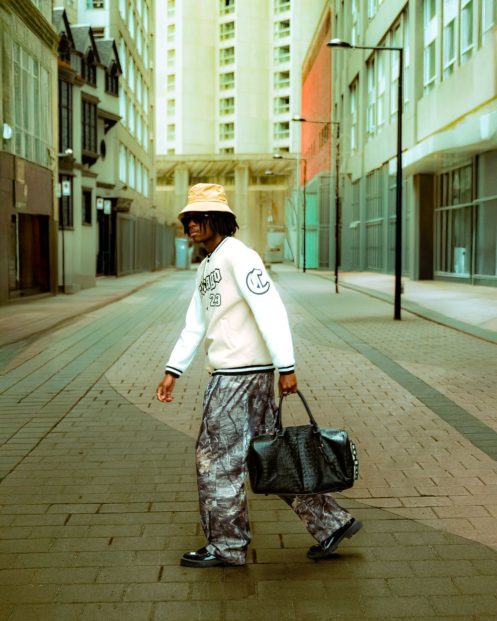 A person in street style clothing carrying a bag walks down an empty urban alleyway between tall buildings.