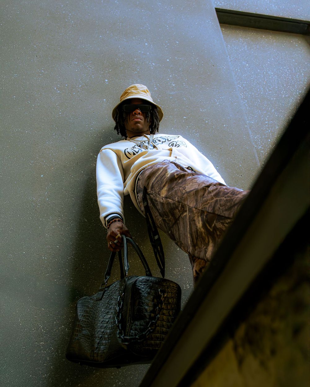 Low-angle view of someone in a white sweater and bucket hat carrying a designer bag on a ledge.