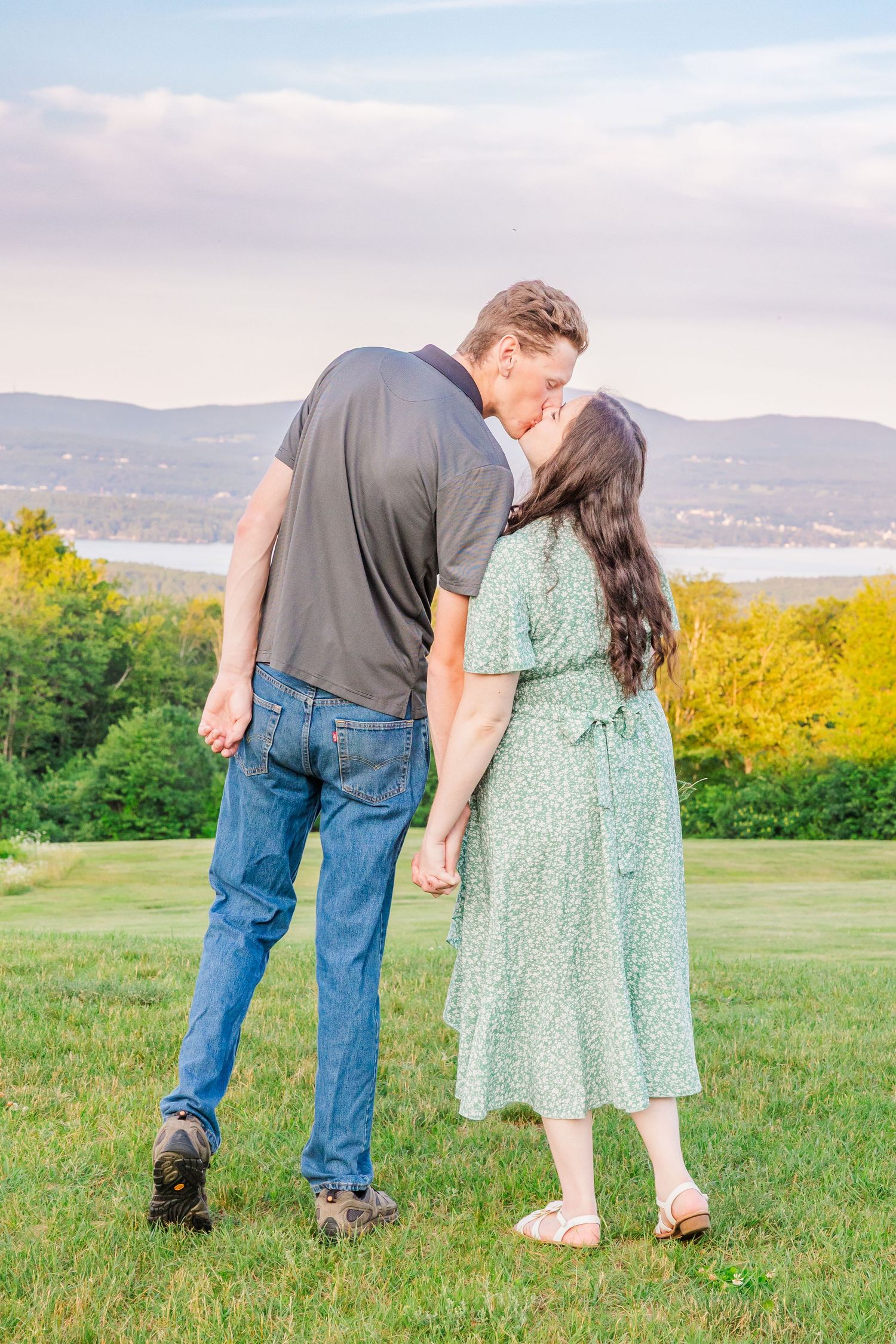 Couple sharing a romantic moment on a grassy hilltop with mountains and water views during golden hour.