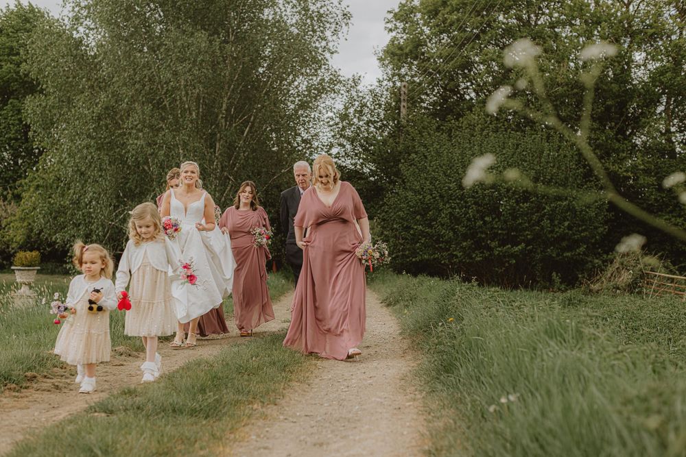 Bridesmaids in pink dresses walk together on a grassy outdoor path.