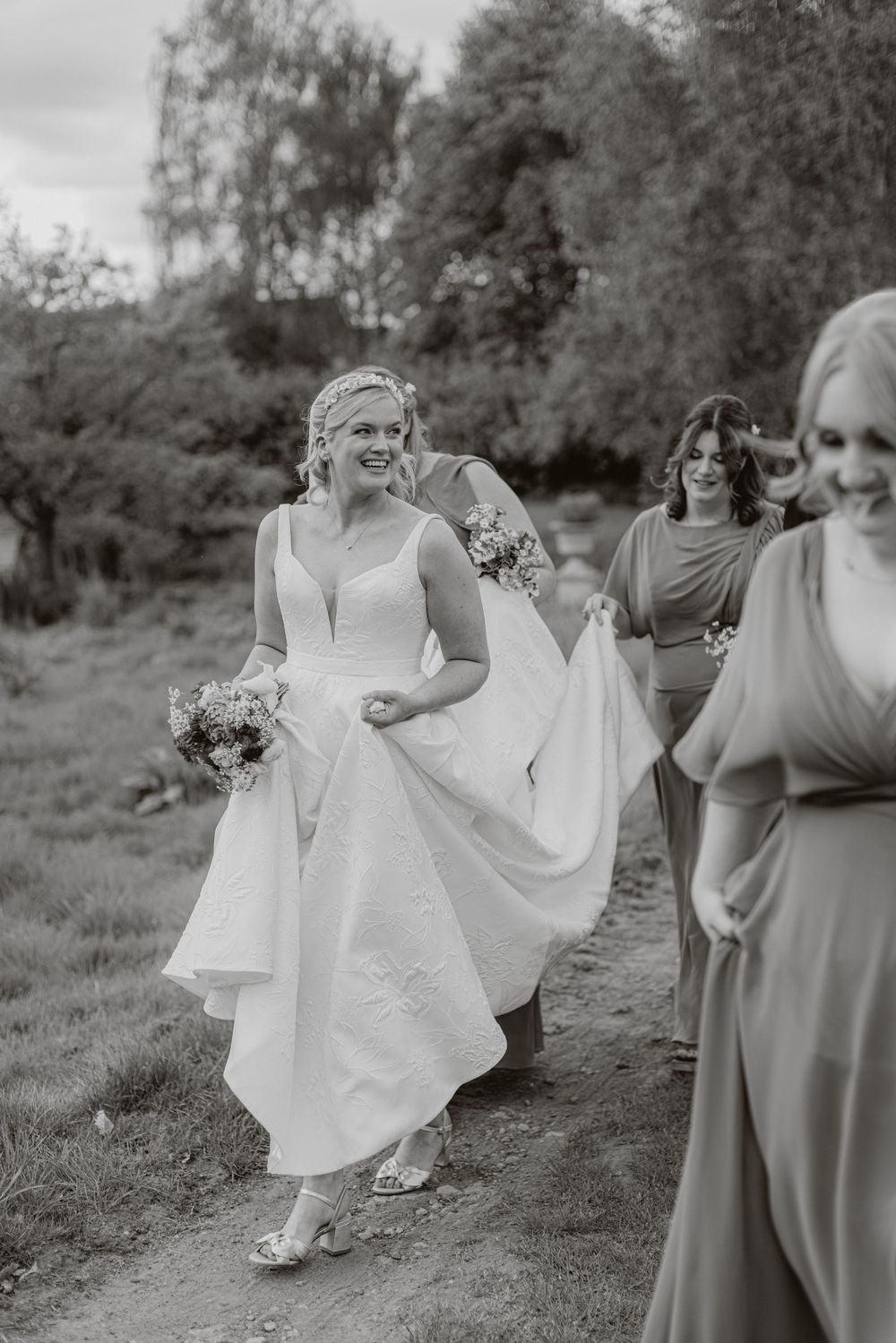 Bridesmaids in flowing dresses walk together holding bouquets on a grassy hillside.