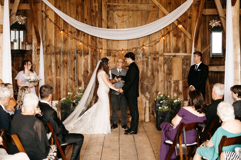A wedding ceremony takes place inside a rustic wooden barn decorated with white fabric draping and floral arrangements.