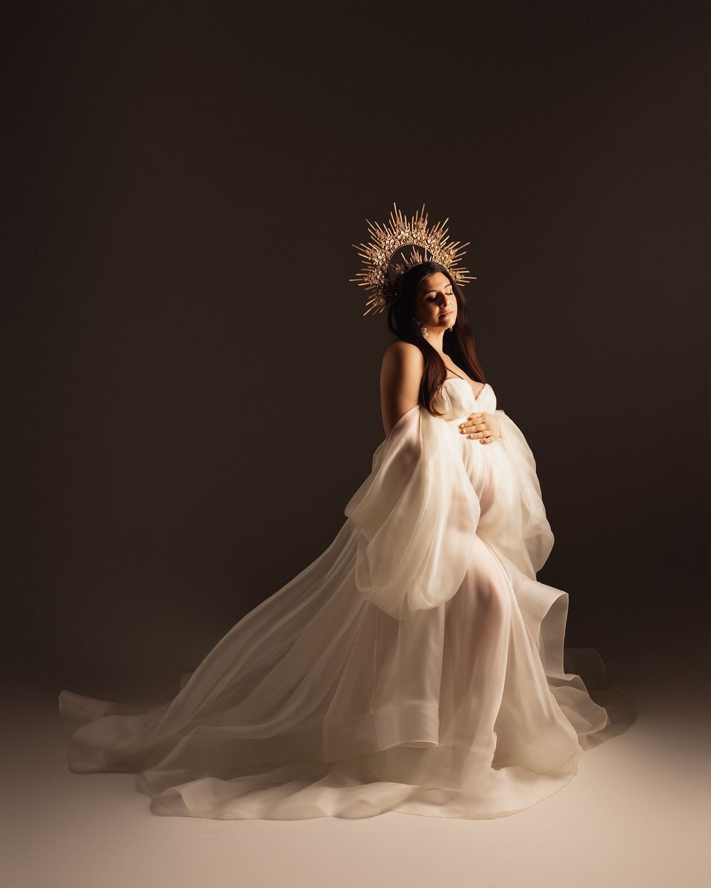 Pregnant woman in white tulle gown and gold crown poses gracefully in a studio maternity portrait.