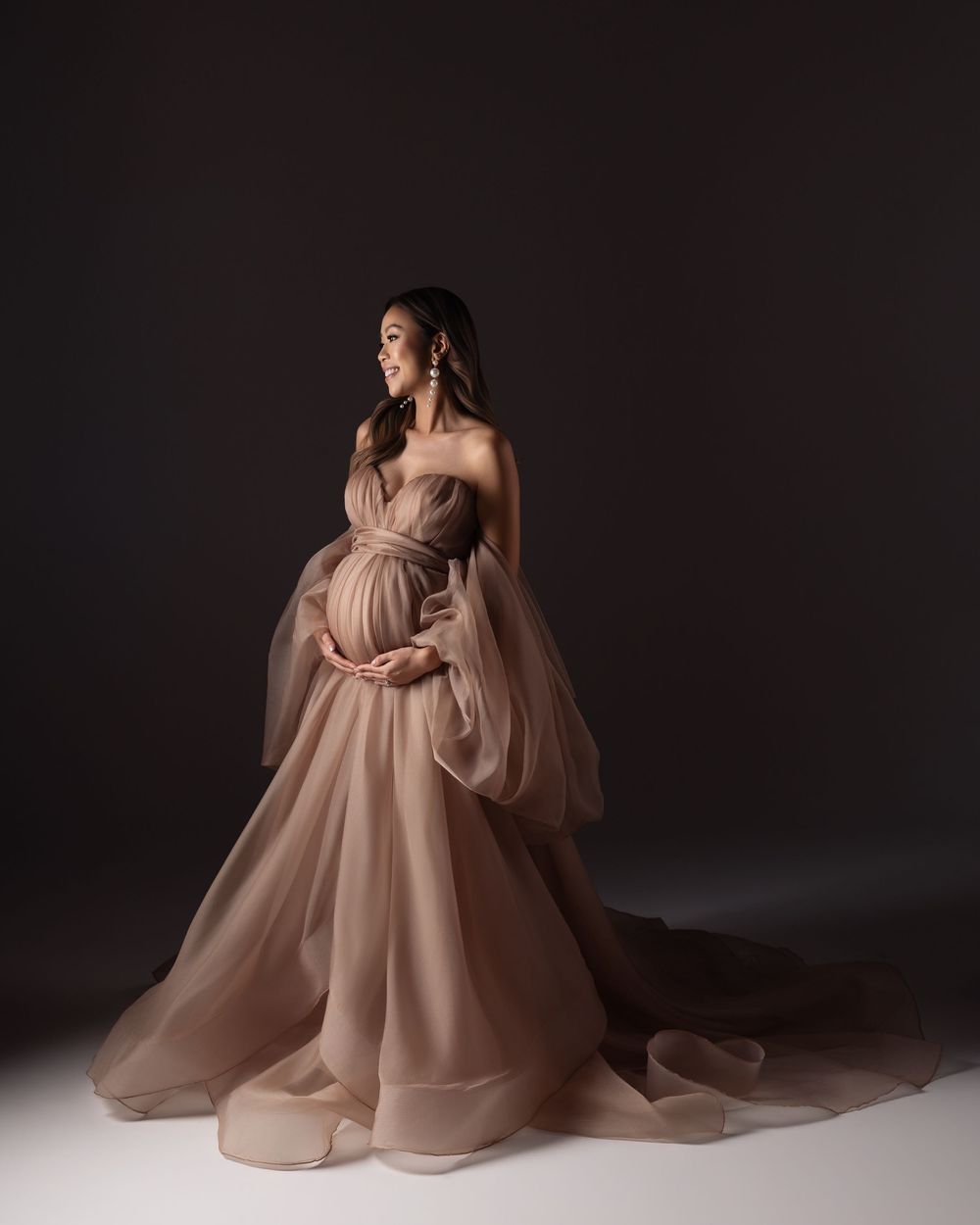 Pregnant woman in elegant dusty rose tulle maternity gown posing gracefully against dark studio background.