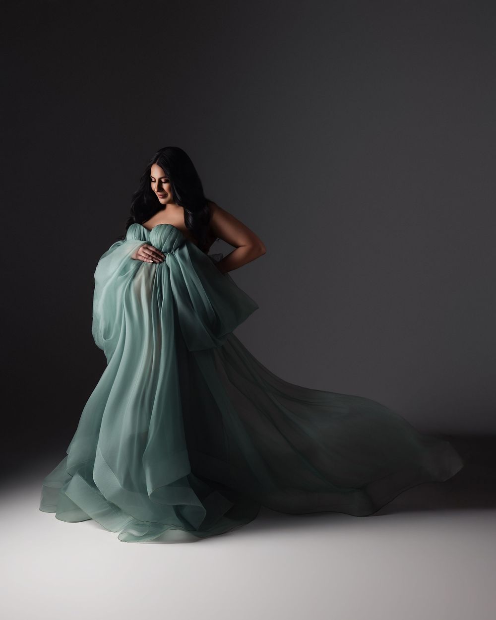 Pregnant woman in flowing sage green tulle maternity gown poses gracefully against dark studio background.