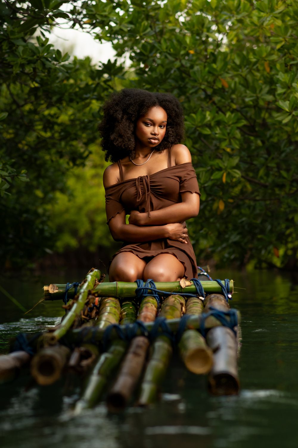 Artistic portrait of someone sitting on bamboo raft in brown dress surrounded by tropical foliage.