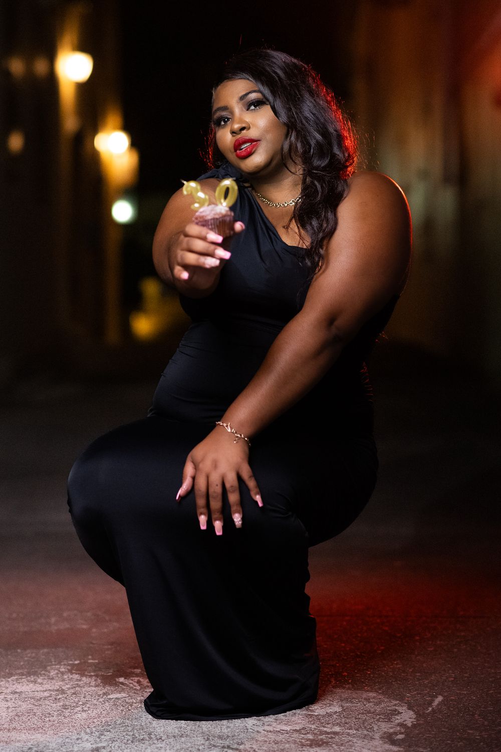 Glamorous nighttime portrait with cocktail in black evening dress under city lights.
