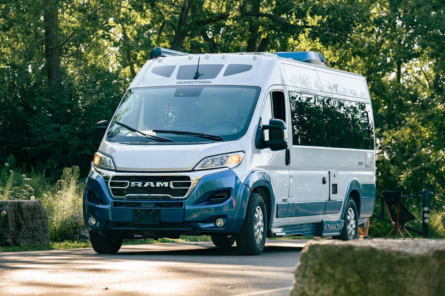 Modern white Ram ProMaster camper van driving on a tree-lined road in a natural setting.