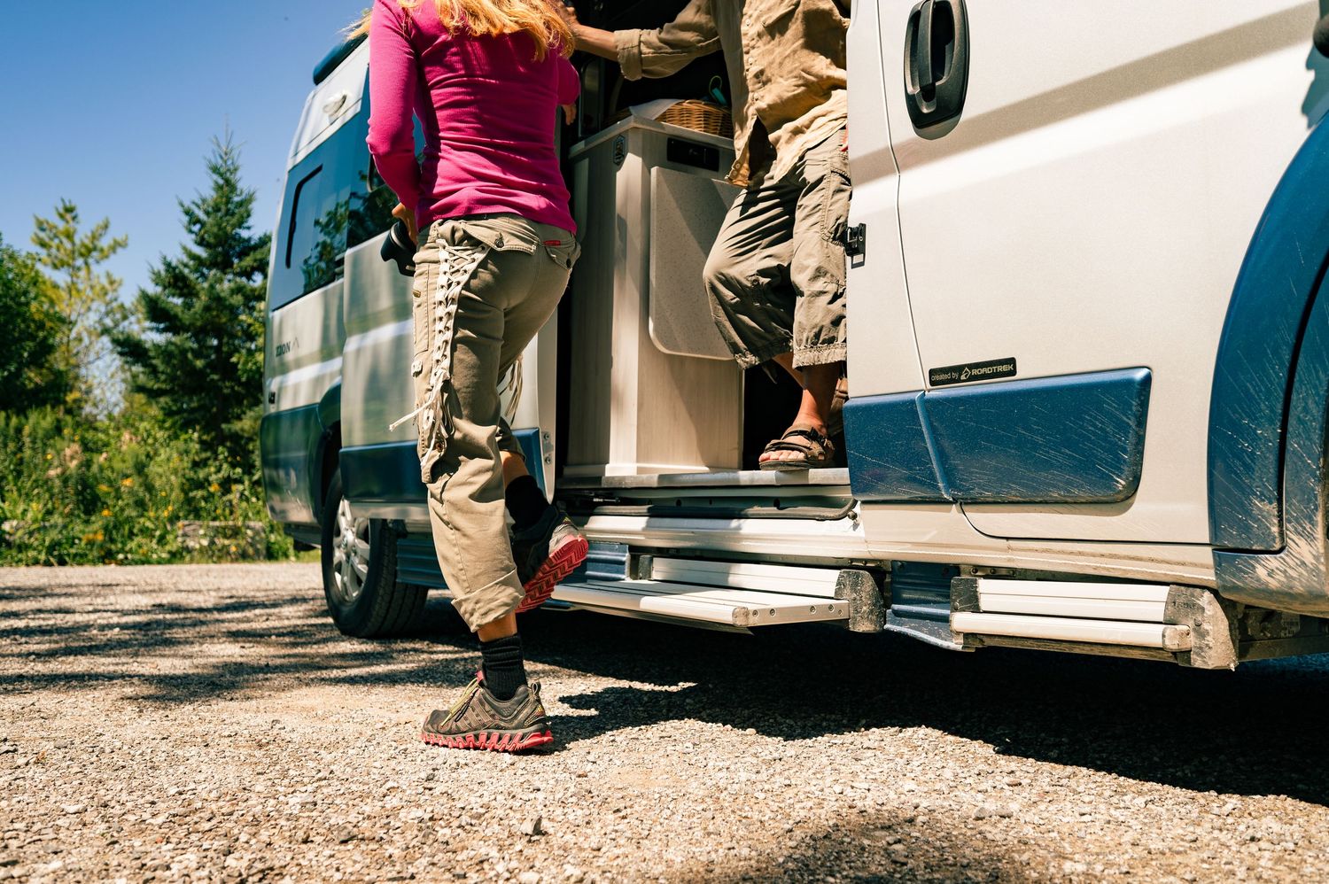 Two people stand on the steps of a white RV parked on a gravel surface with trees visible in the background.