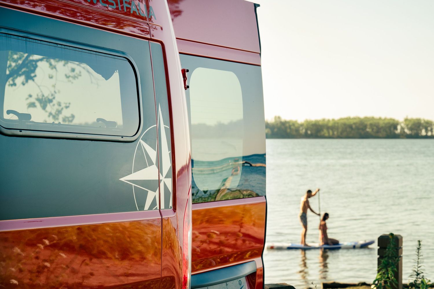 Orange van parked near a lake with reflective sunset views and silhouettes of paddleboarders in the distance.