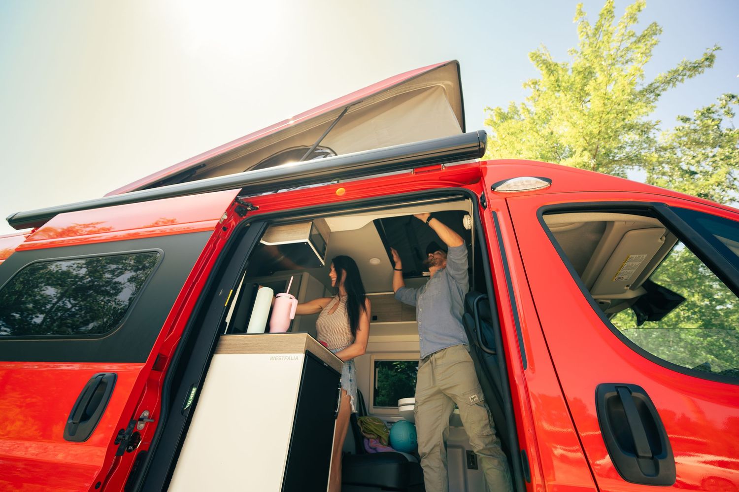 A camping van interior shows someone organizing storage compartments in a bright red conversion vehicle.