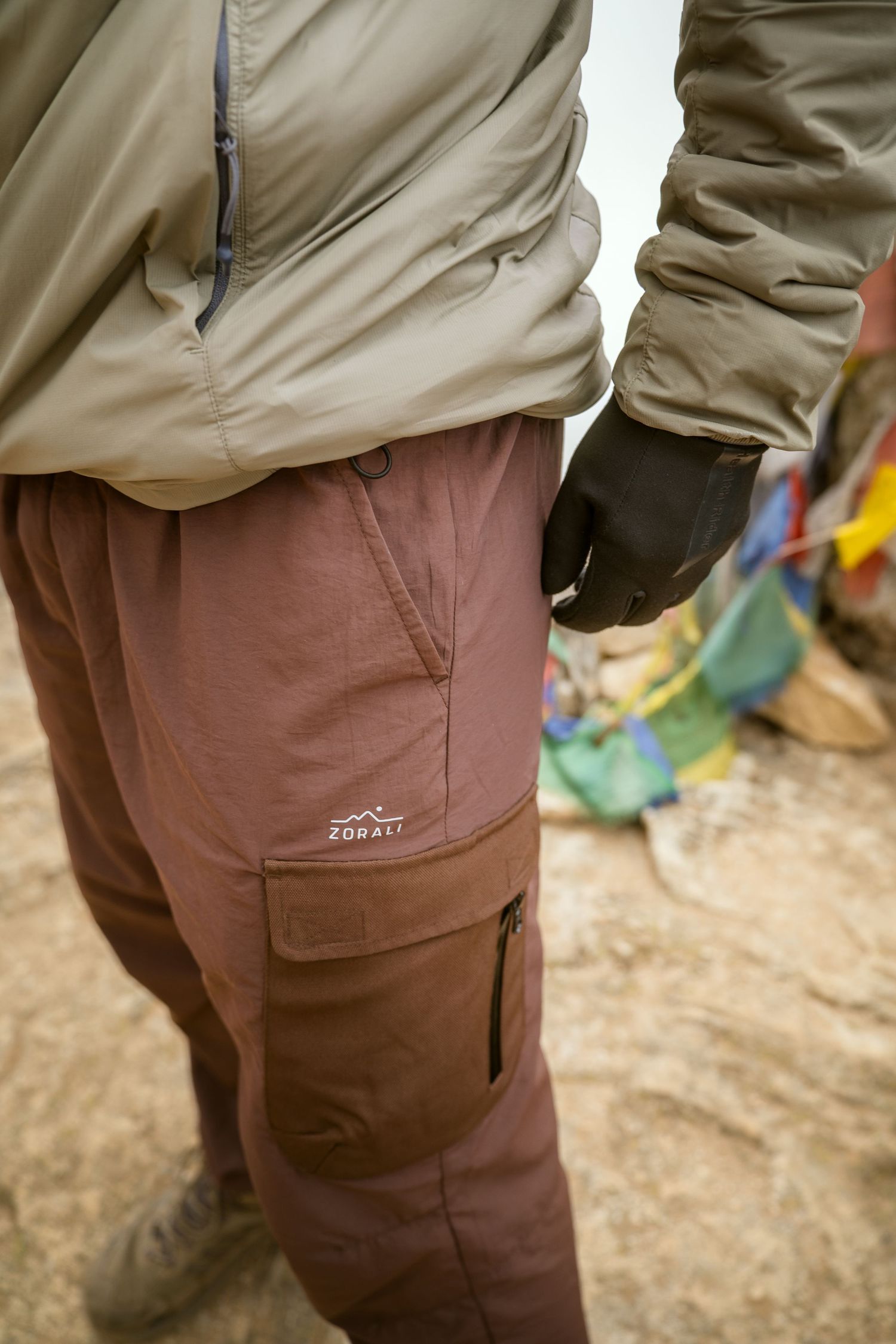 Close-up of khaki outdoor cargo pants showing a branded patch pocket against a dirt ground.