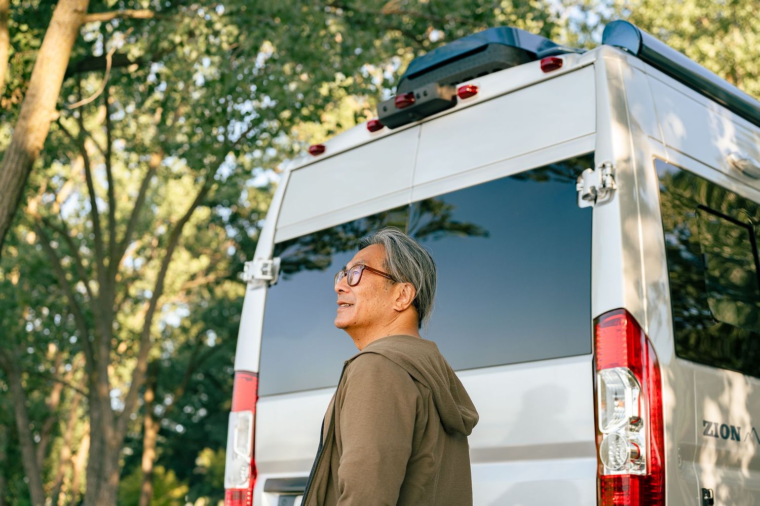 Middle-aged Asian man with gray hair and glasses stands beside a white camper van surrounded by lush green trees.