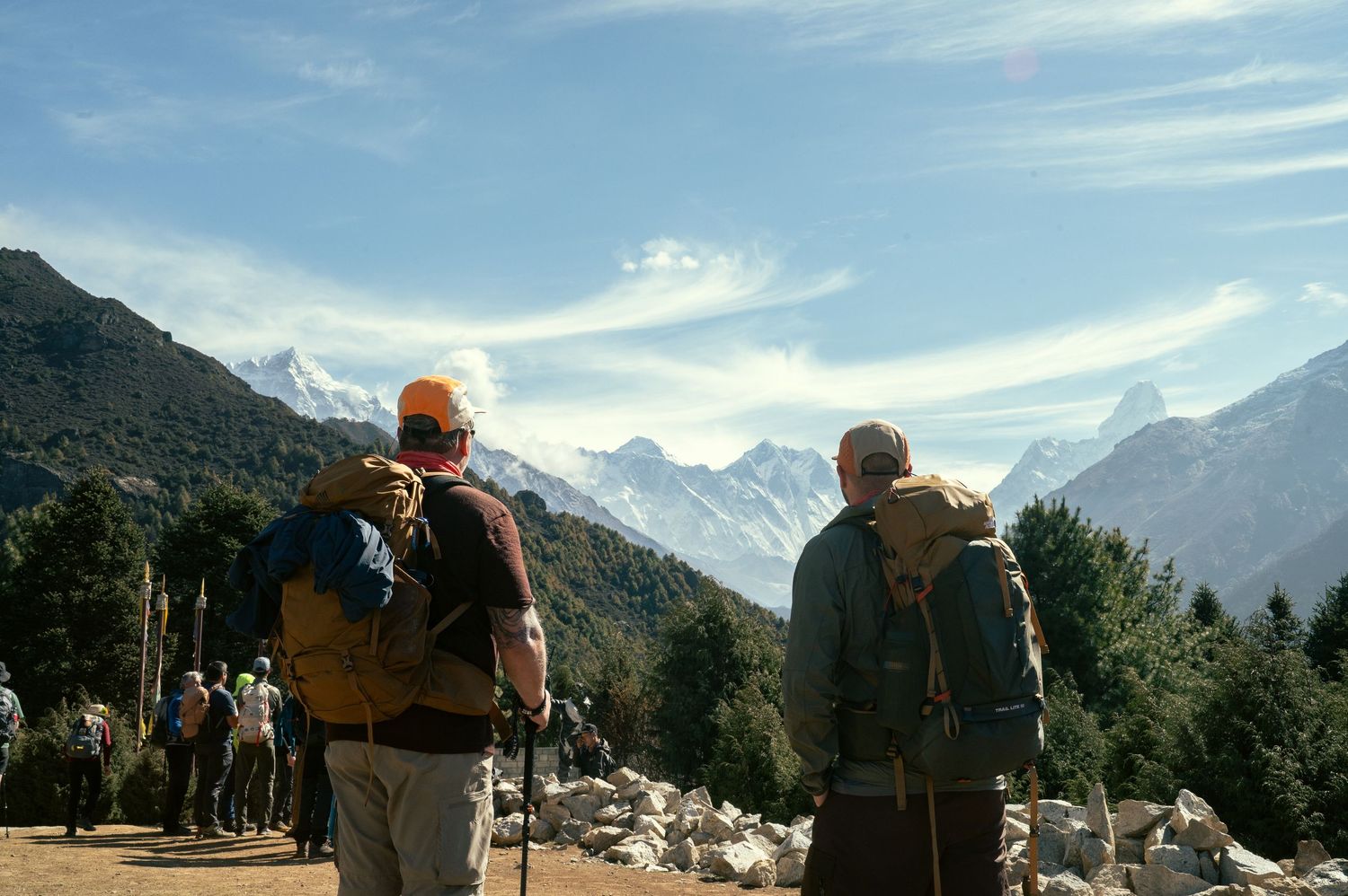 Hikers with backpacks stand on mountain trail overlooking snowy peaks and valley in scenic wilderness landscape.