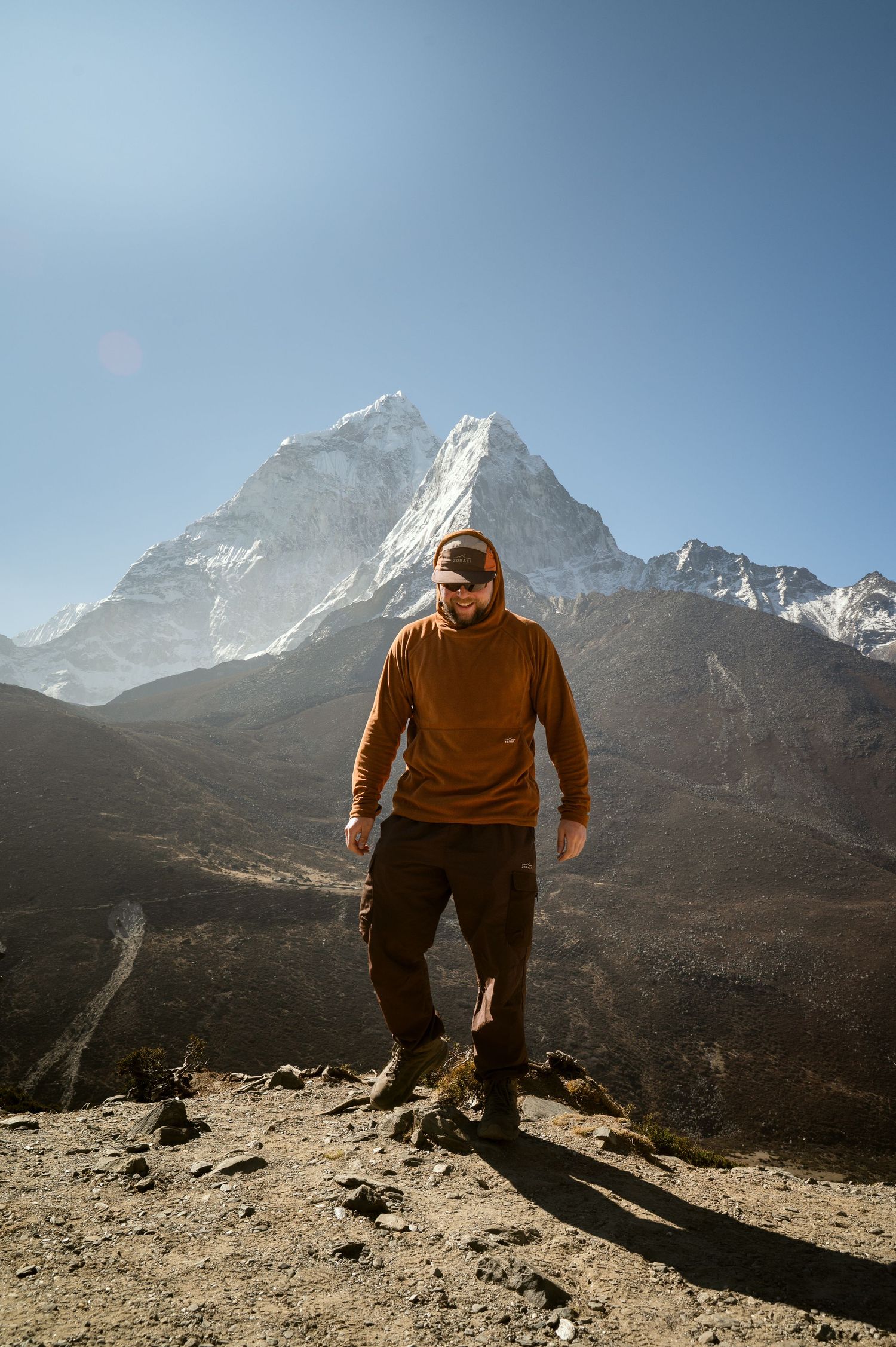 A hiker in a brown sweater stands on a rocky path against the backdrop of a majestic snow-capped Himalayan mountain peak.