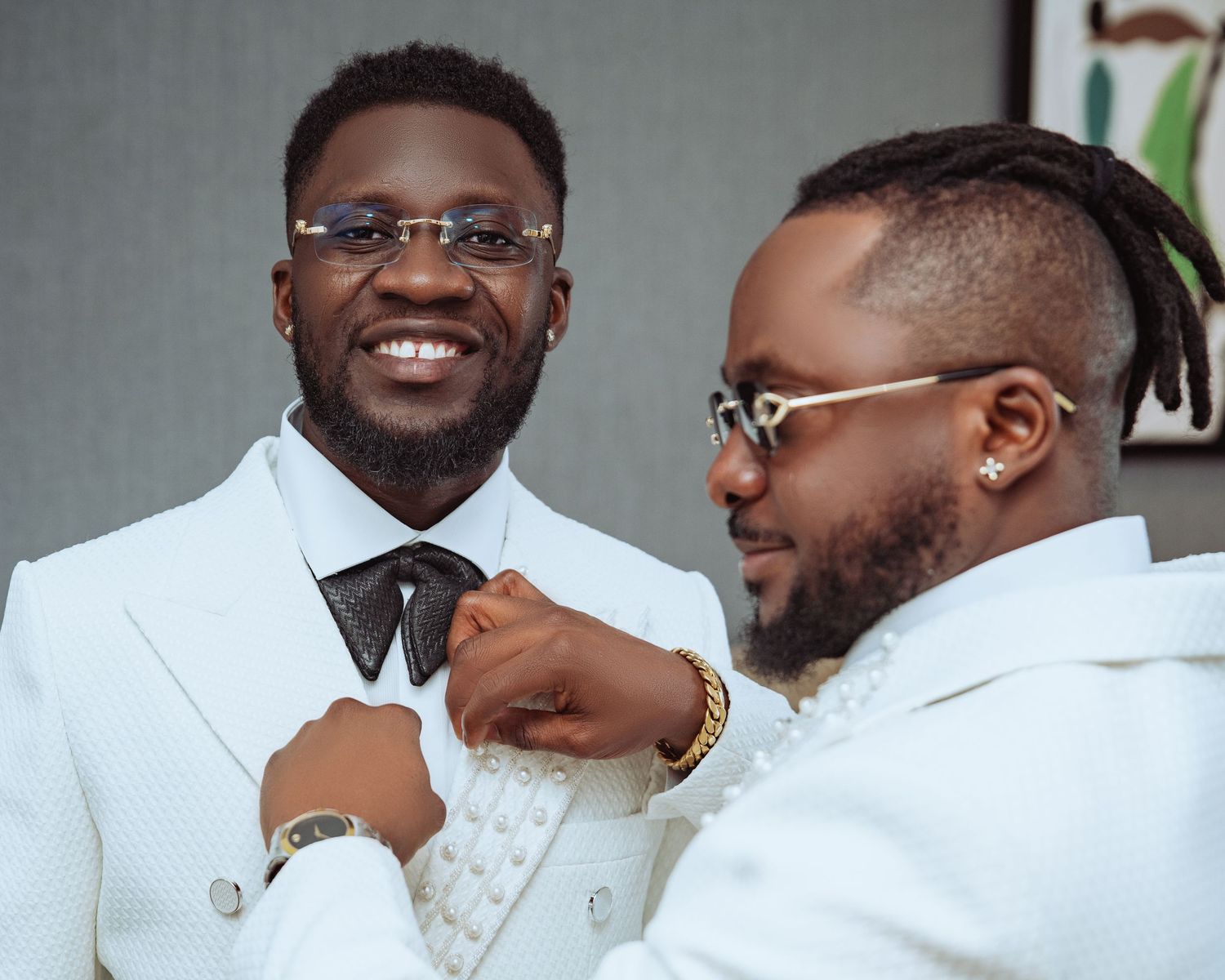 Two people in white suits adjusting each other's formal attire while smiling.